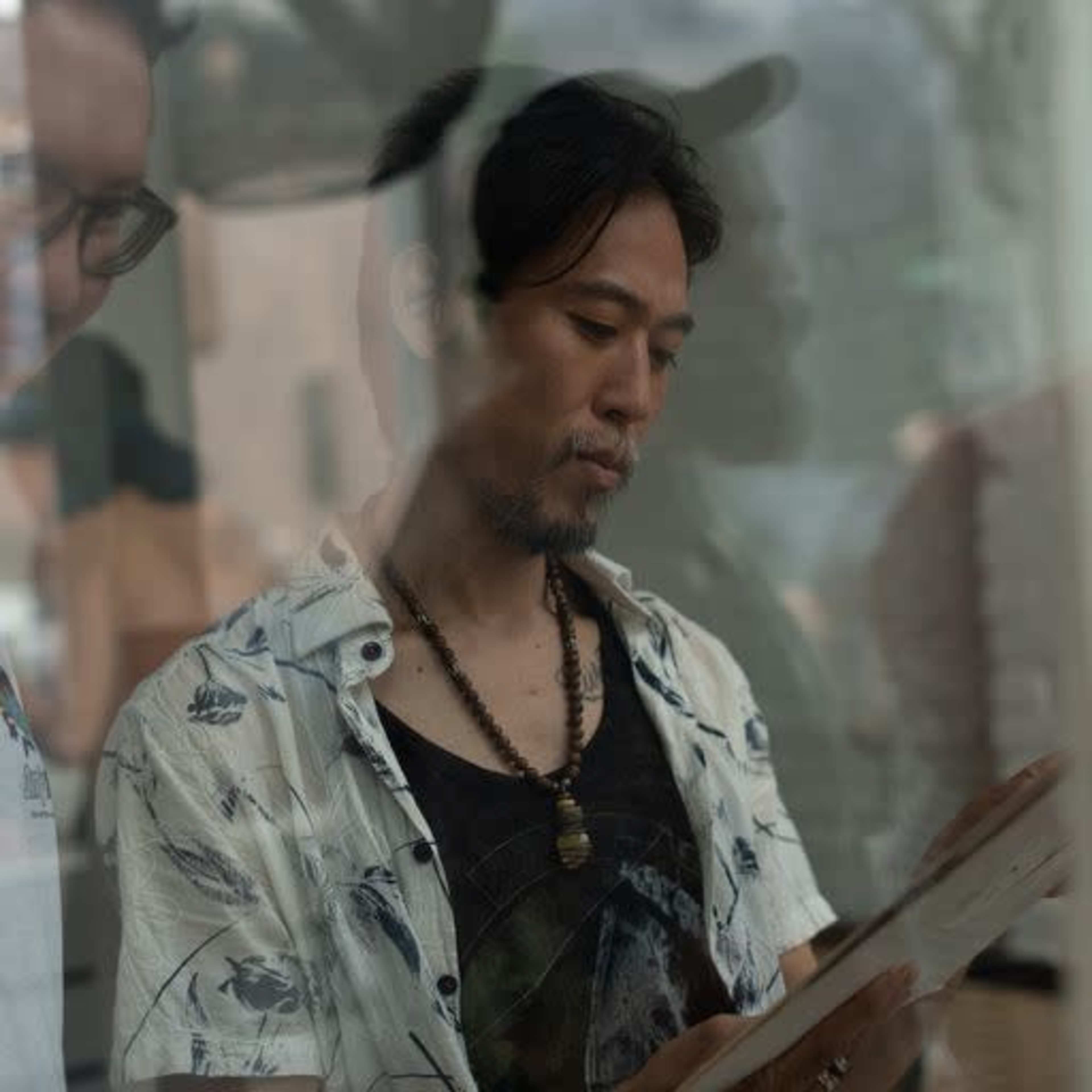 Two men are standing closely together, with one man wearing a patterned shirt and examining a piece of paper while the other looks on.