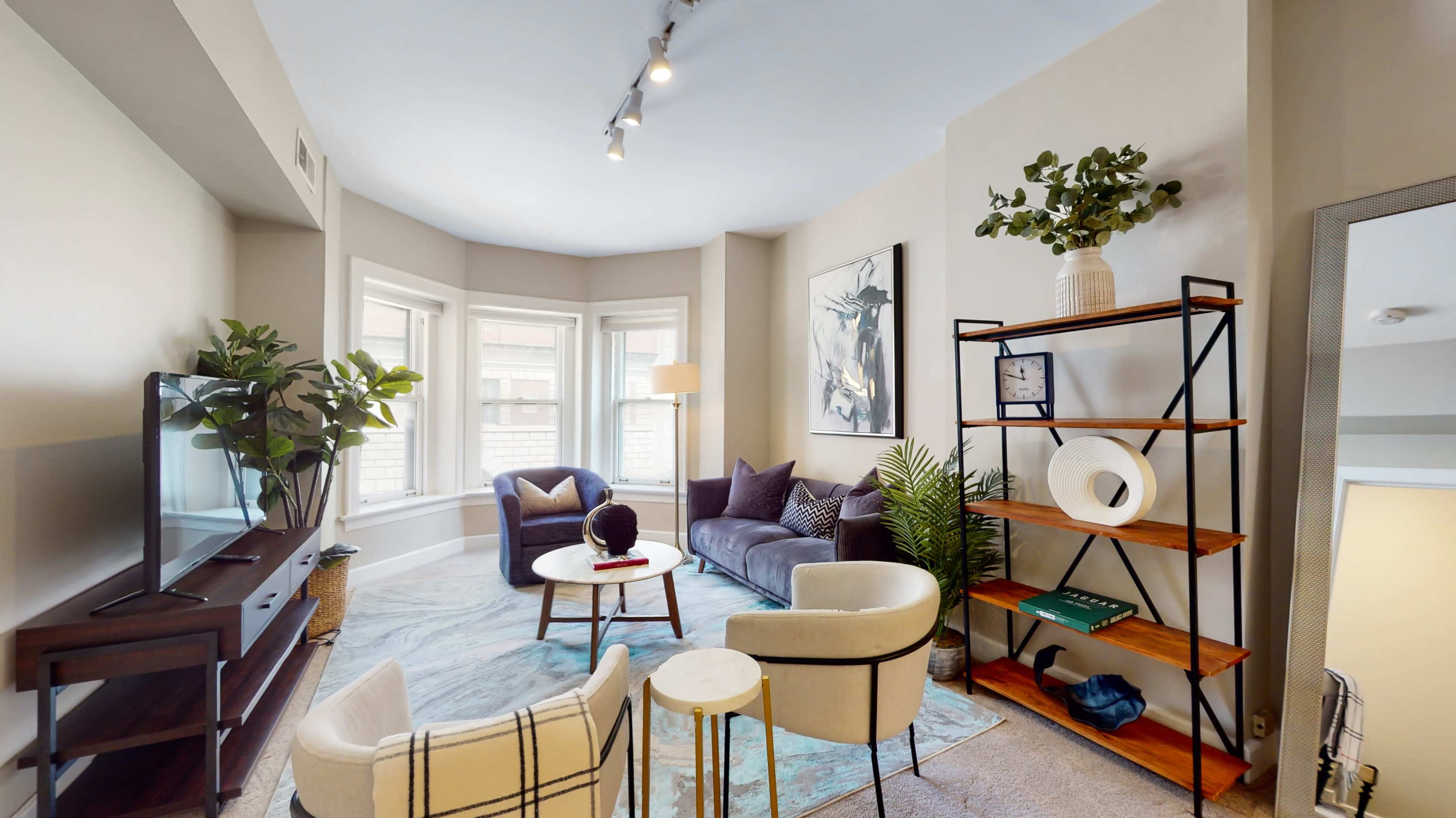 The image shows a cozy living room featuring a sofa, armchairs, a coffee table, a television, and decorative plants in a sunlit space.
