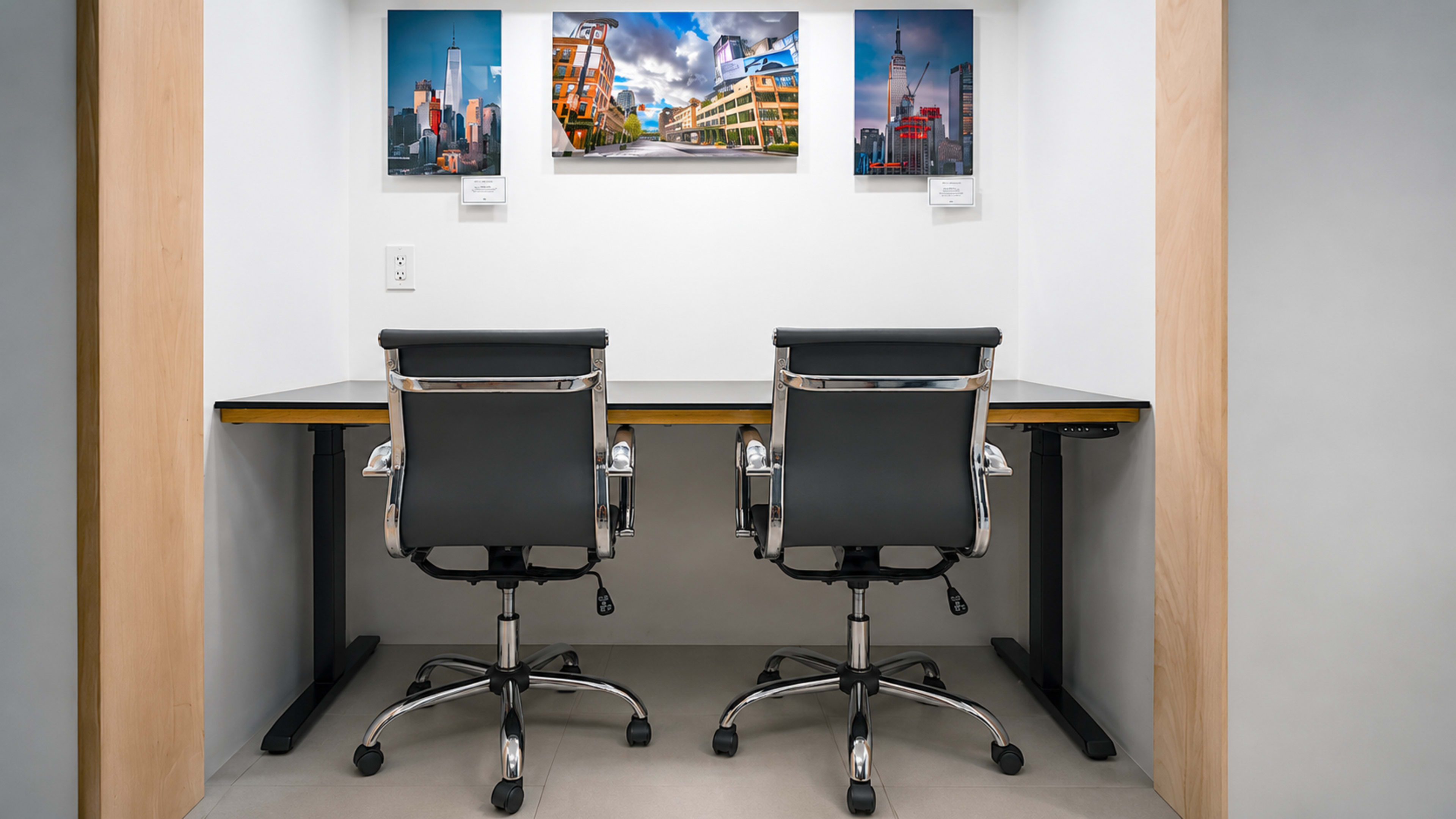 The image shows a small workspace featuring a modern desk with two black office chairs and three framed photographs on the wall above.