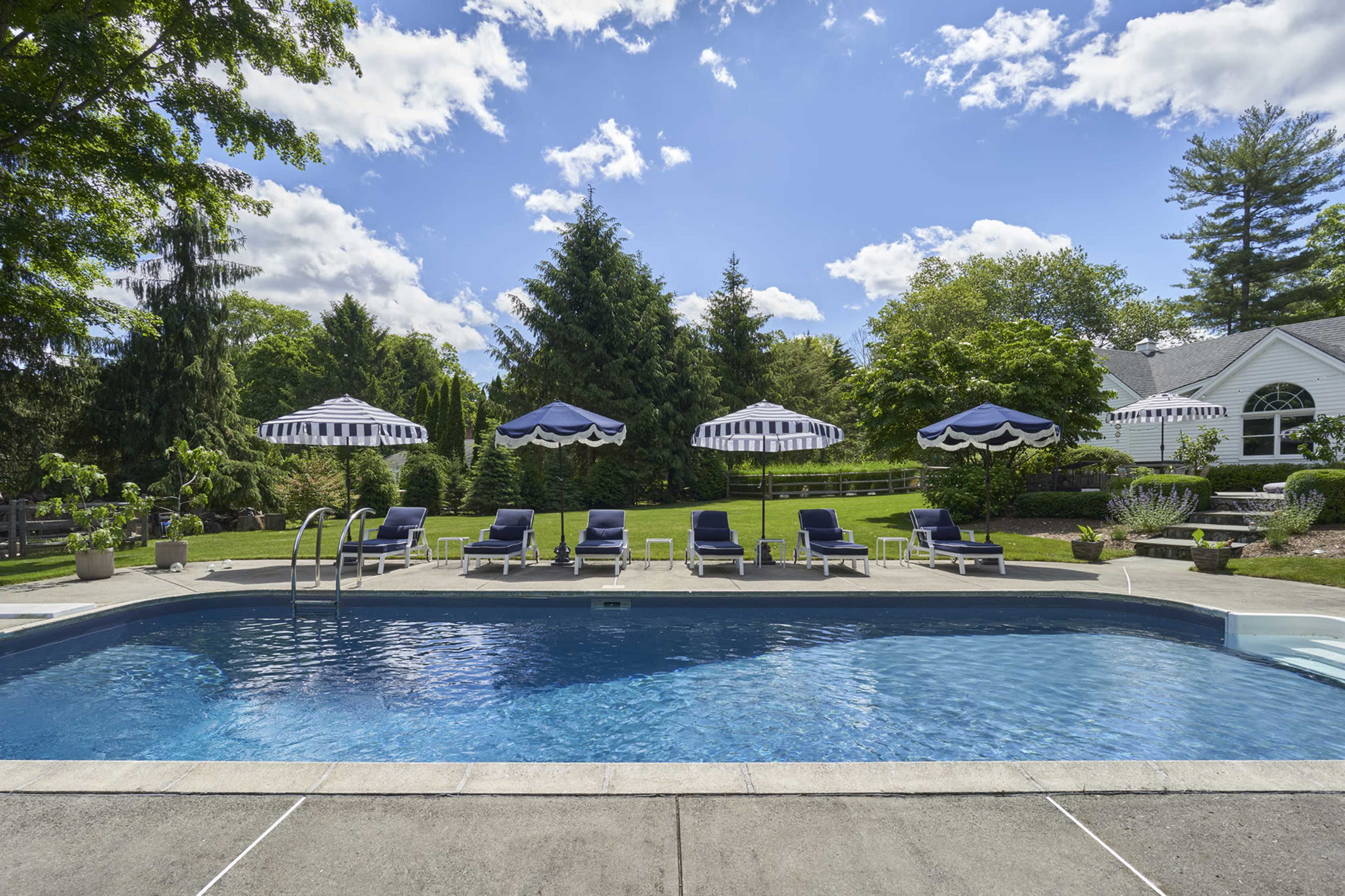 A clear swimming pool is surrounded by deck chairs and umbrellas in a landscaped backyard under a blue sky.
