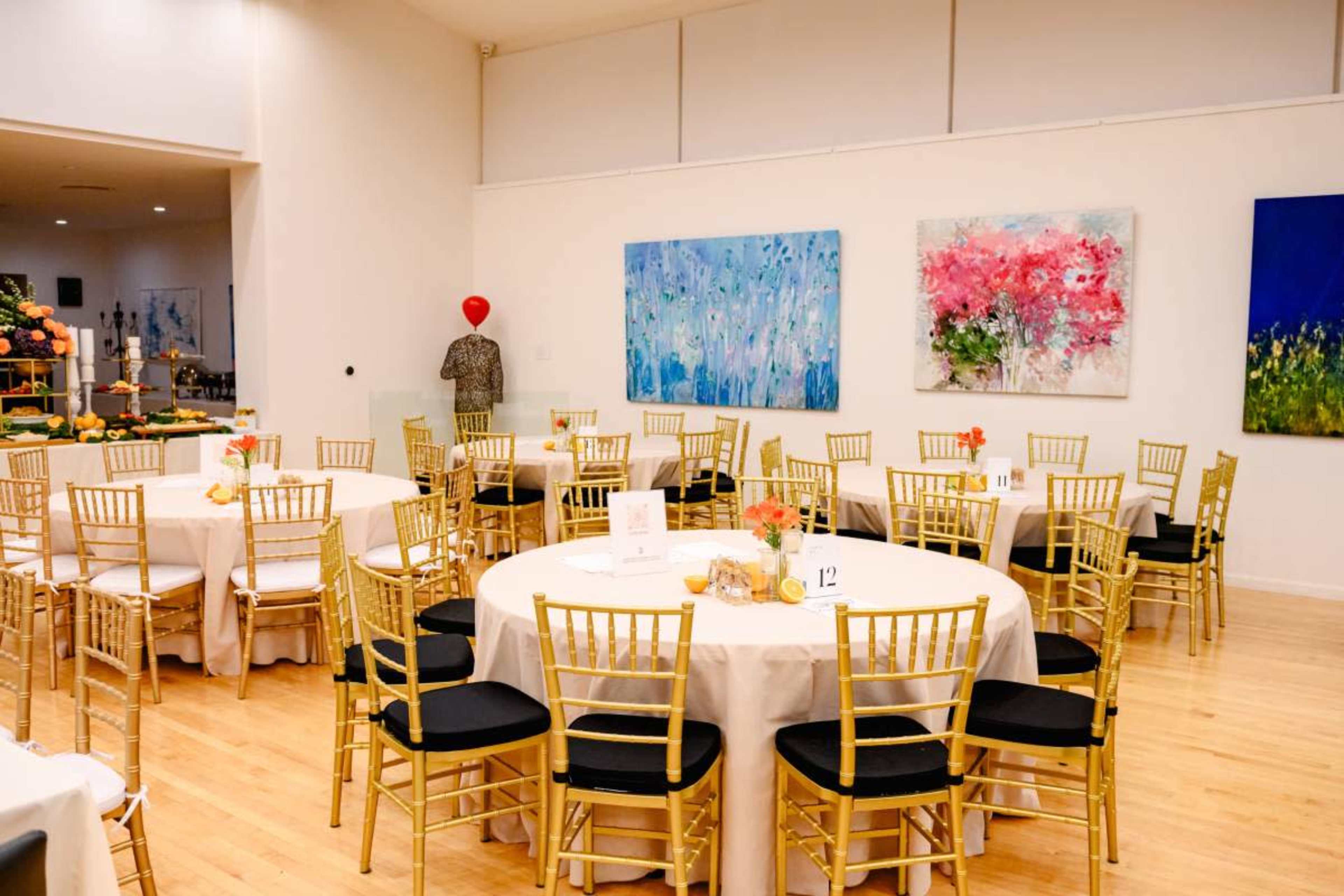 A spacious event room features round tables with white tablecloths and gold chairs, adorned with floral centerpieces and colorful artwork on the walls.