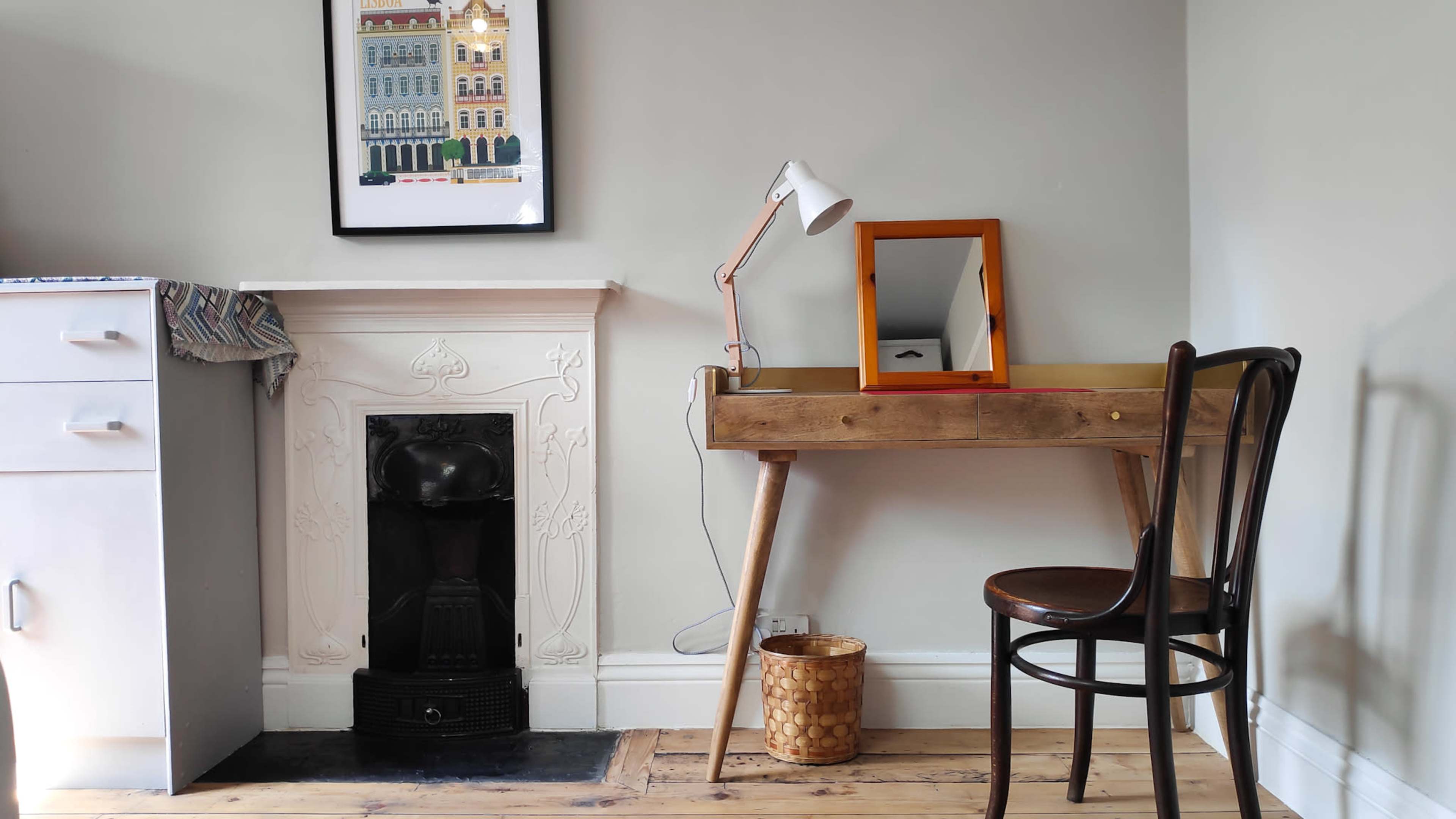 A wooden desk with a lamp and a mirror sits next to a decorative fireplace in a room with light gray walls and wooden flooring.