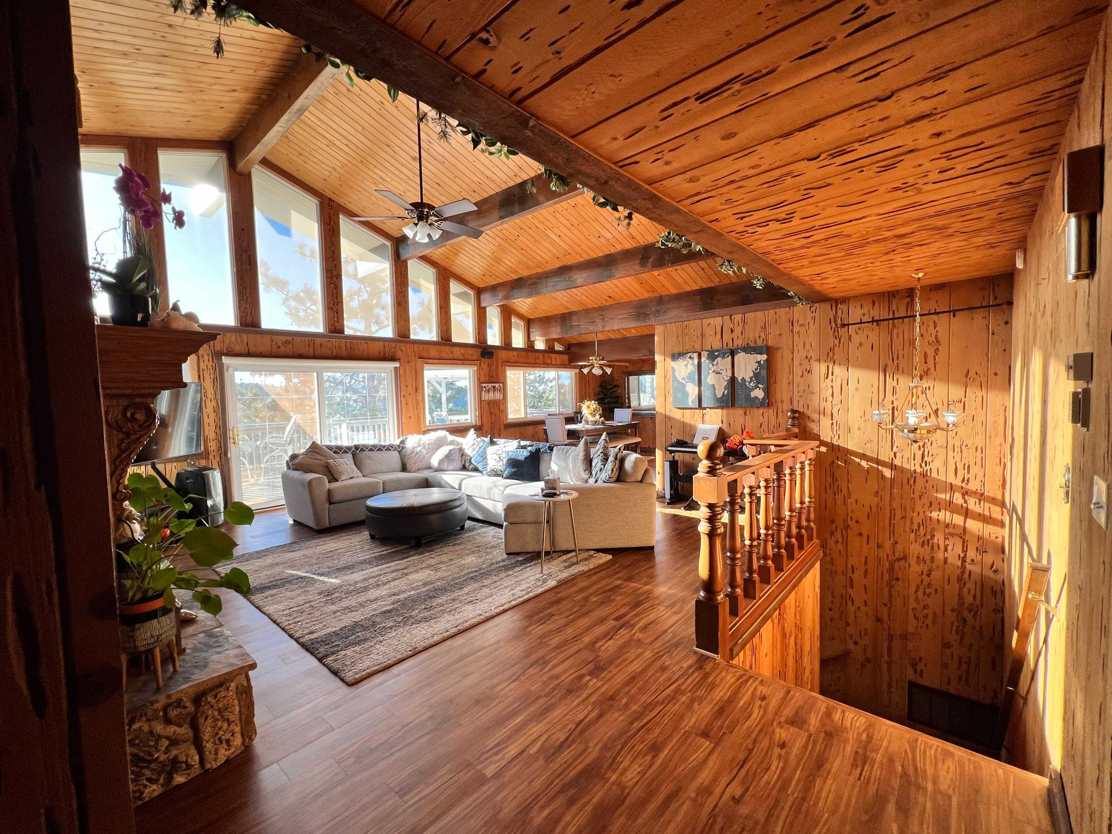 A spacious living room with wooden walls and ceiling, large windows allowing natural light, and a cozy seating area featuring a sectional sofa and coffee table.
