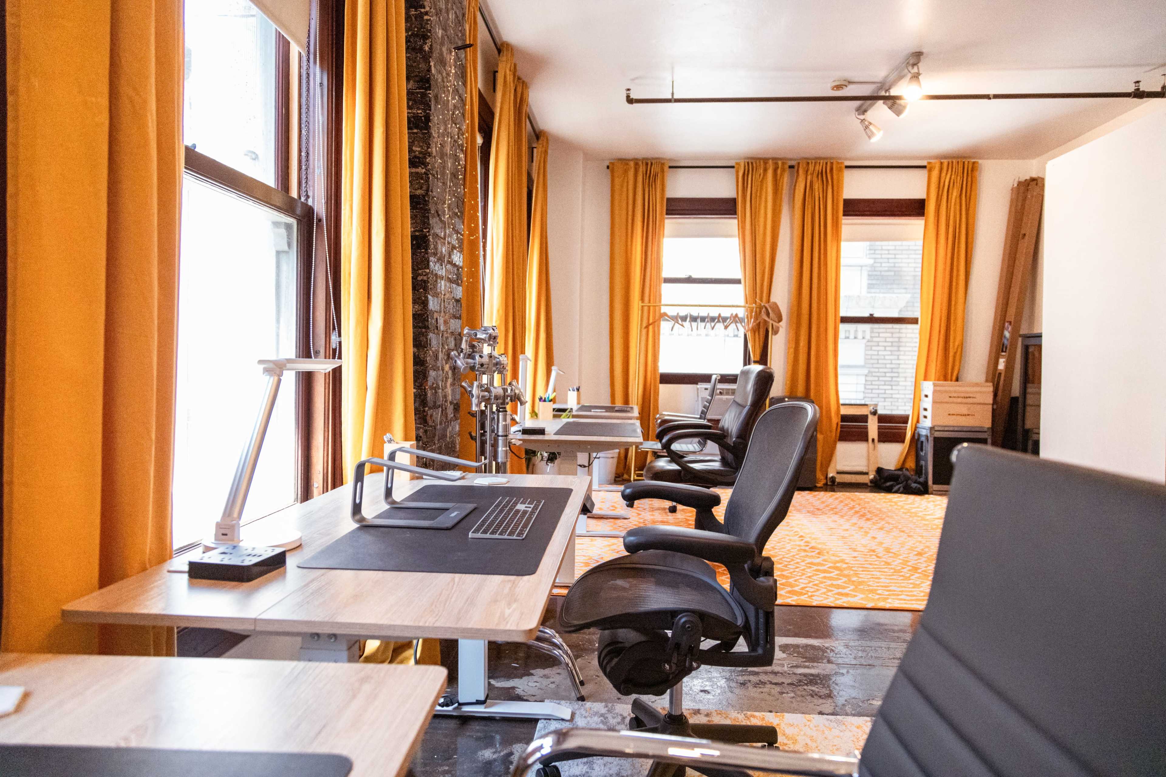 The image shows a modern office space with several desks, ergonomic chairs, and large windows dressed with orange curtains.