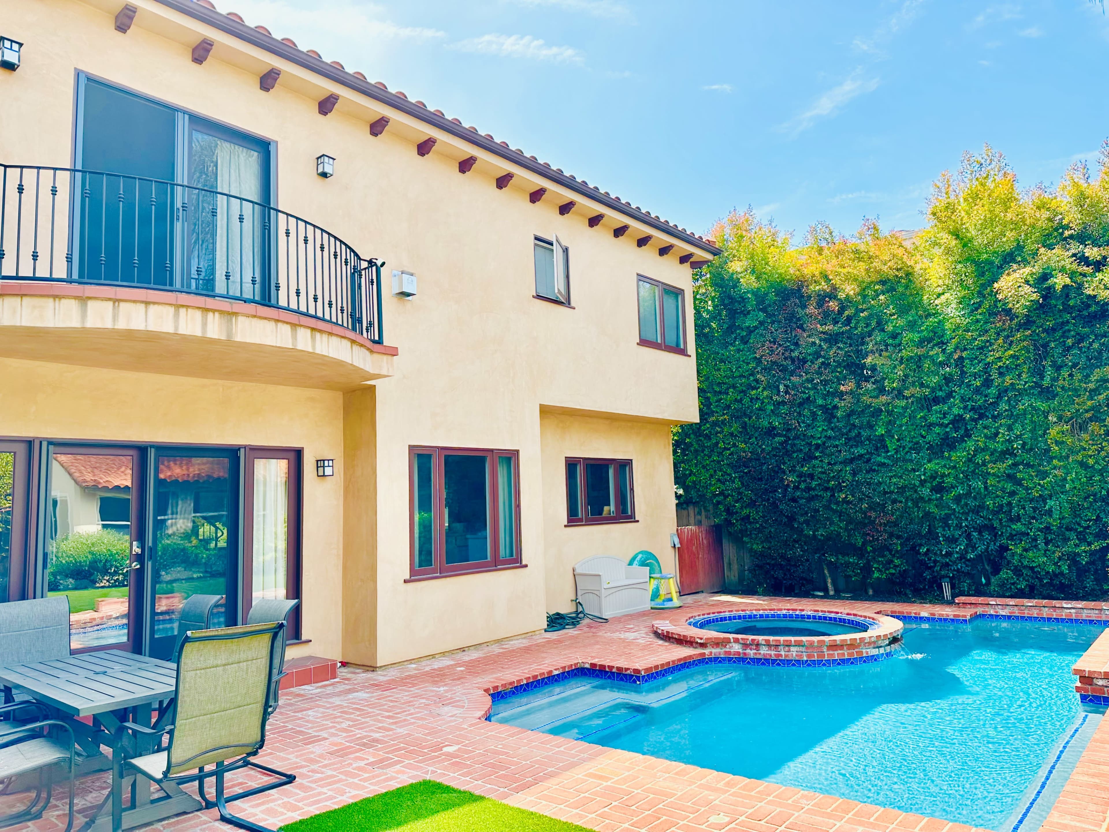 A two-story house with a balcony overlooks a backyard pool and spa surrounded by greenery.