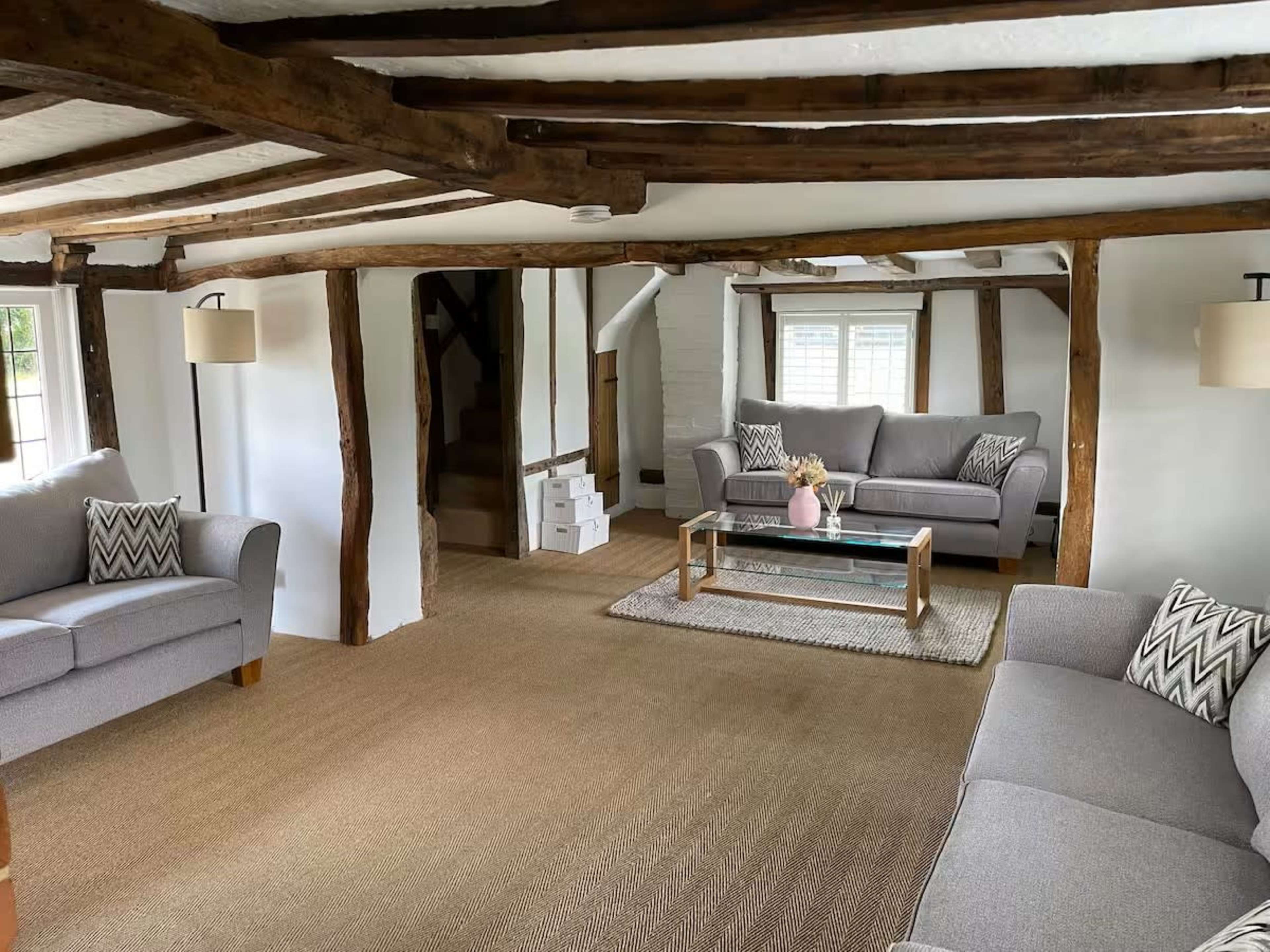 The image shows a cozy living room with exposed wooden beams, two gray sofas, a glass coffee table, and natural fiber flooring.