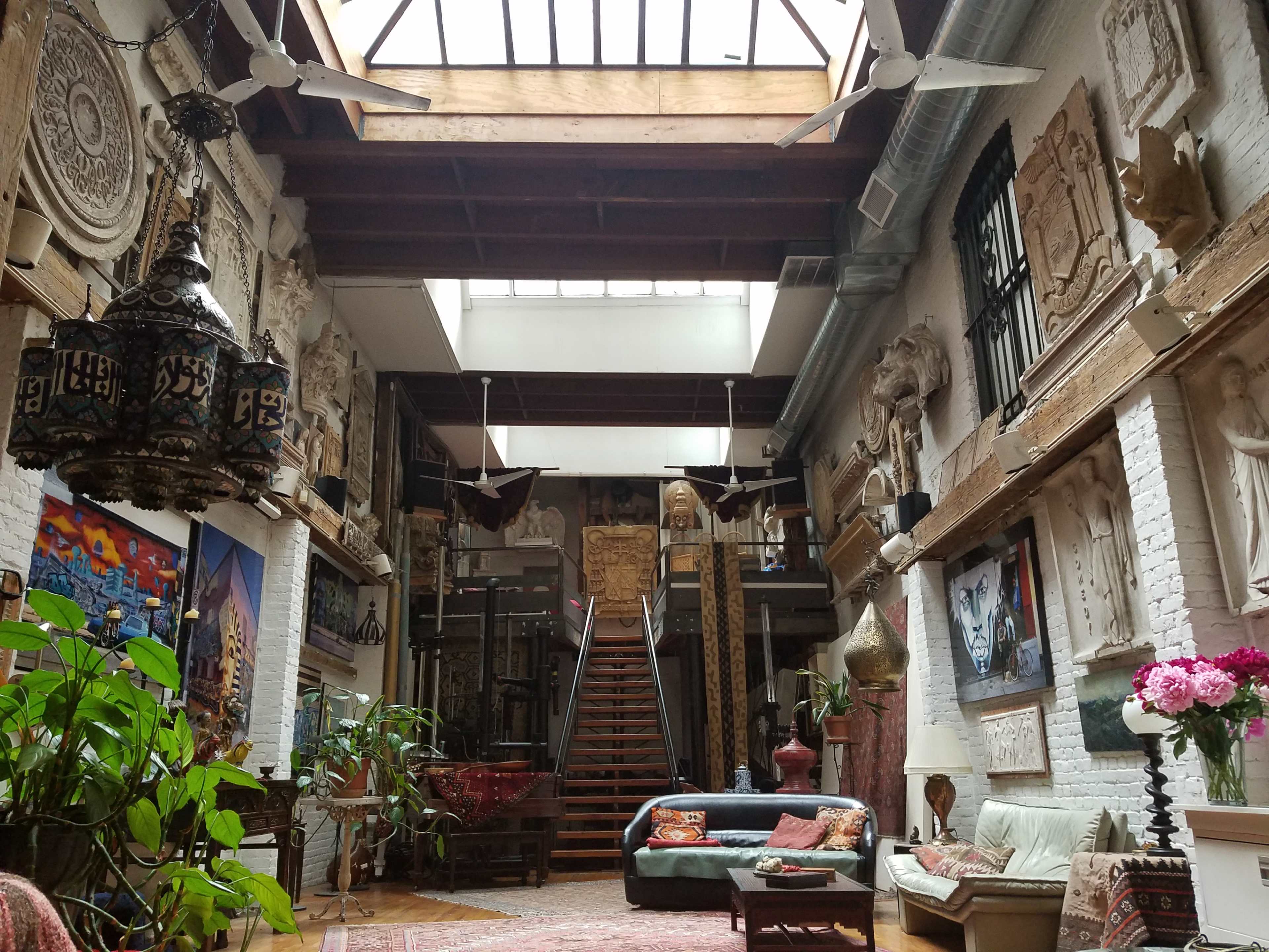 A spacious interior of an art-filled loft with high ceilings, ornate sculptures on the walls, and a staircase leading to an upper level.