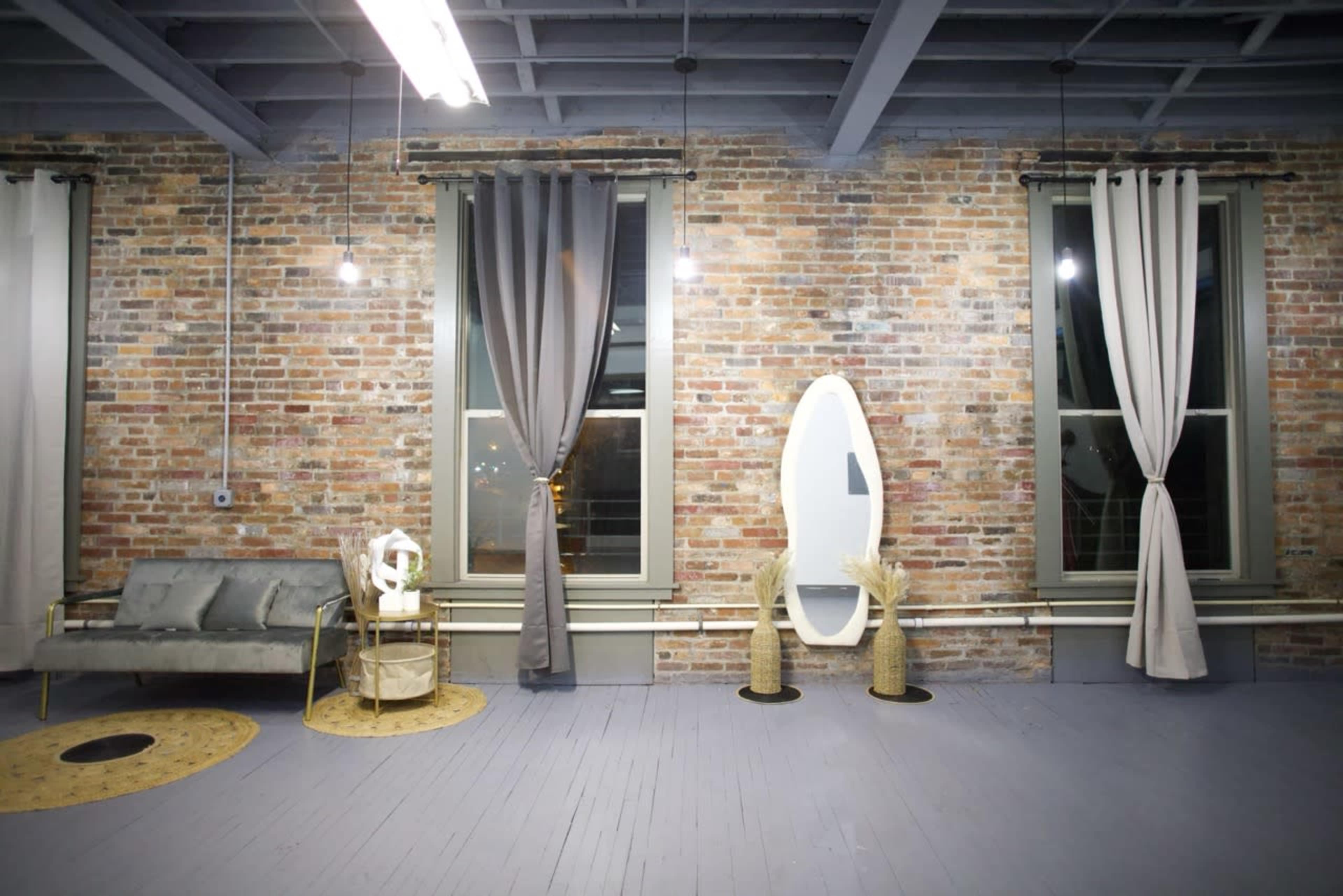The image shows a stylish interior space featuring exposed brick walls, large windows with curtains, a mirror, and modern furniture arranged on a light-colored floor.