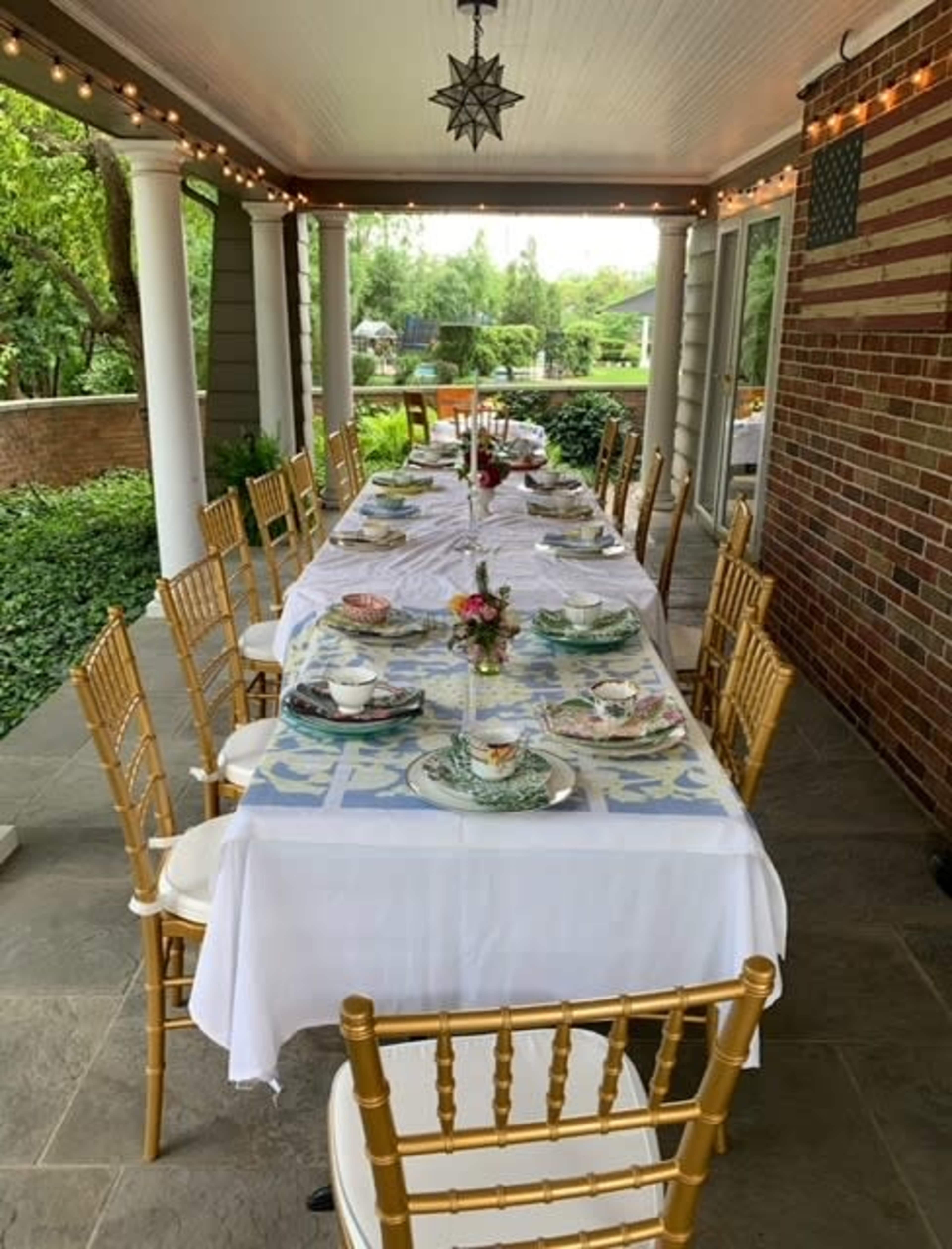 A long dining table is set for a meal on a covered patio, surrounded by greenery and decorated with dishes and floral centerpieces.