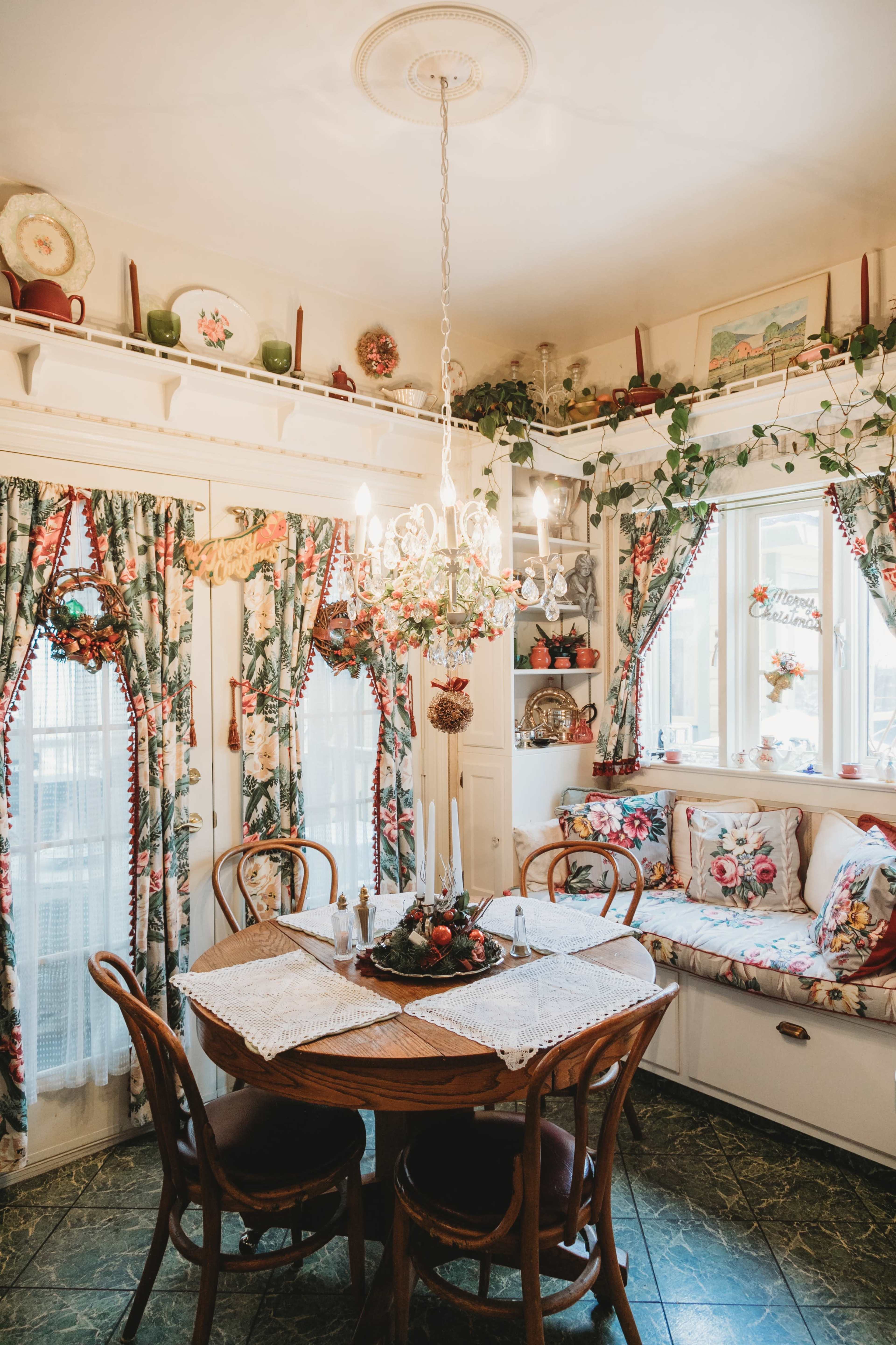 The image shows a quaint kitchen with a round wooden table covered by a lace tablecloth, surrounded by four wooden chairs and adorned with floral curtains and decorative items on the shelves and window sills.