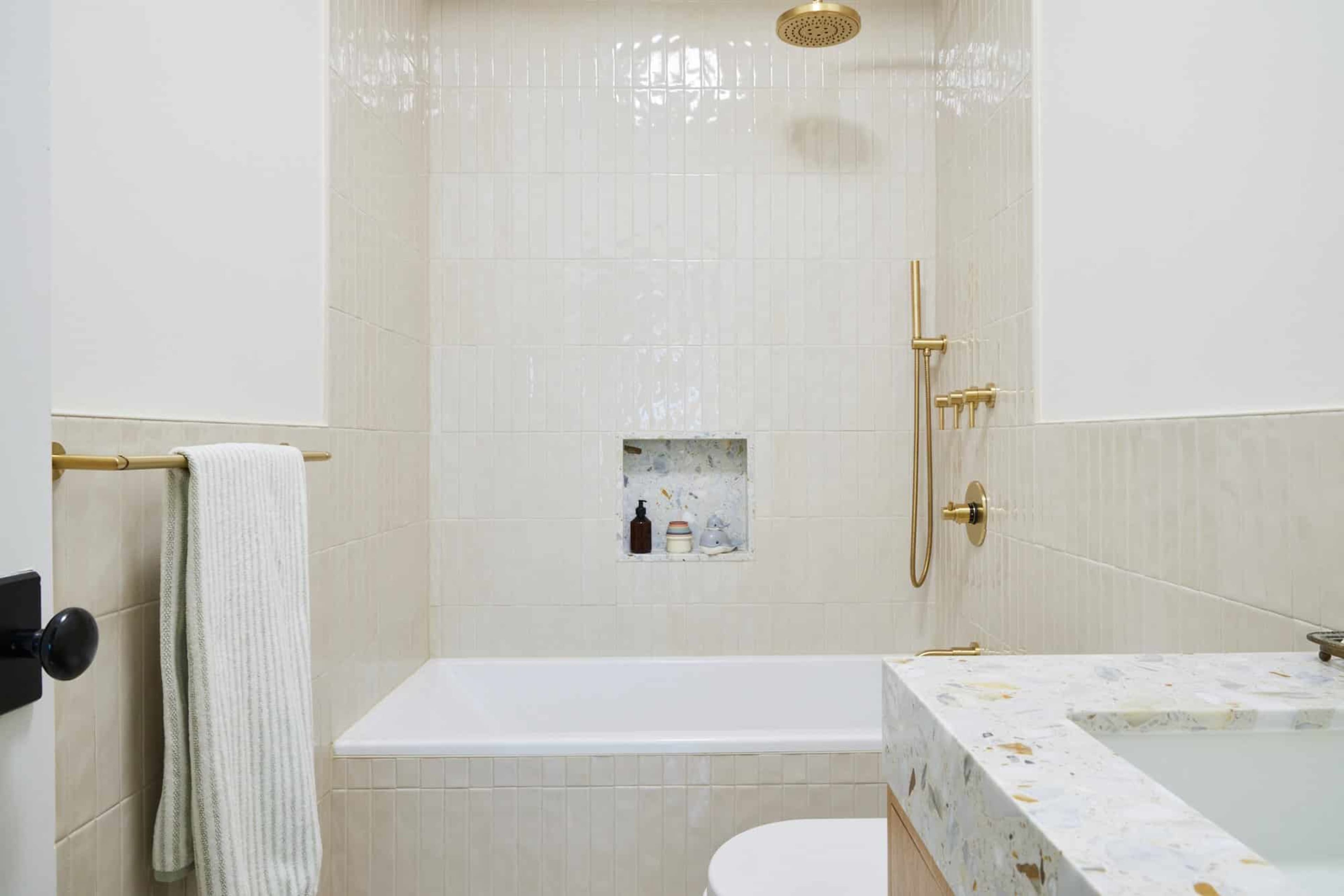 The image depicts a modern bathroom featuring a white bathtub, a gold shower fixture, and a niche with decorative items set into tiled walls.