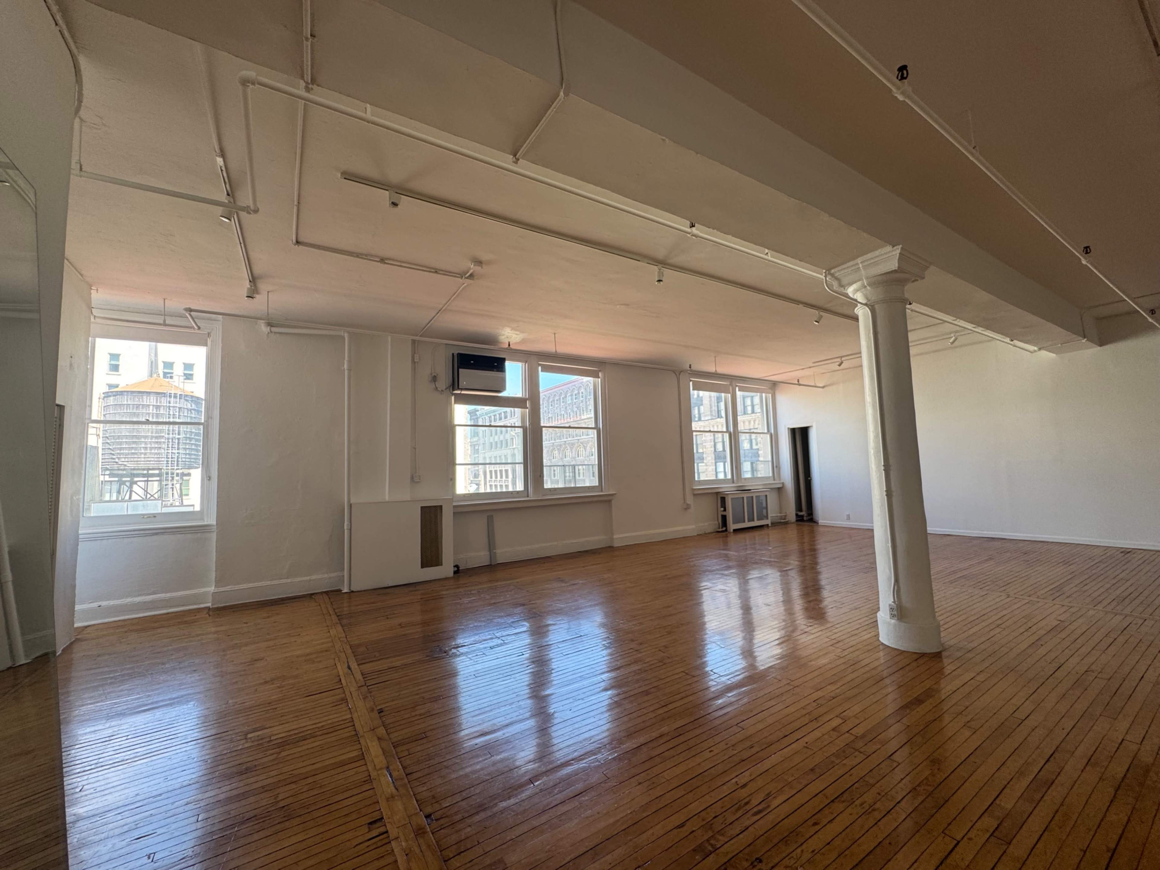 The image shows a spacious, empty room with hardwood floors, large windows, and white walls.