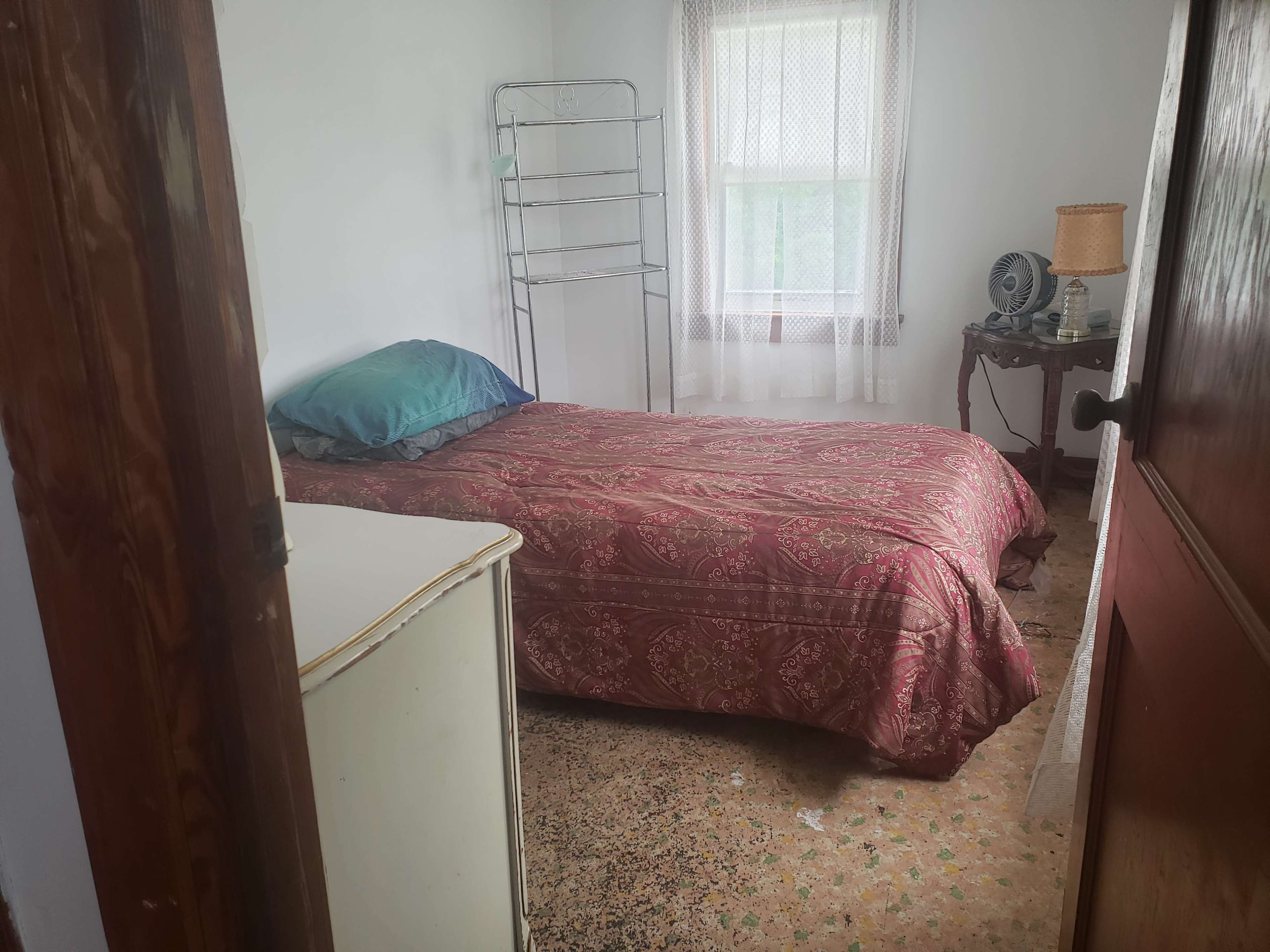 The room features a single bed with a red patterned quilt, a small bedside table, and a white dresser near an open door.