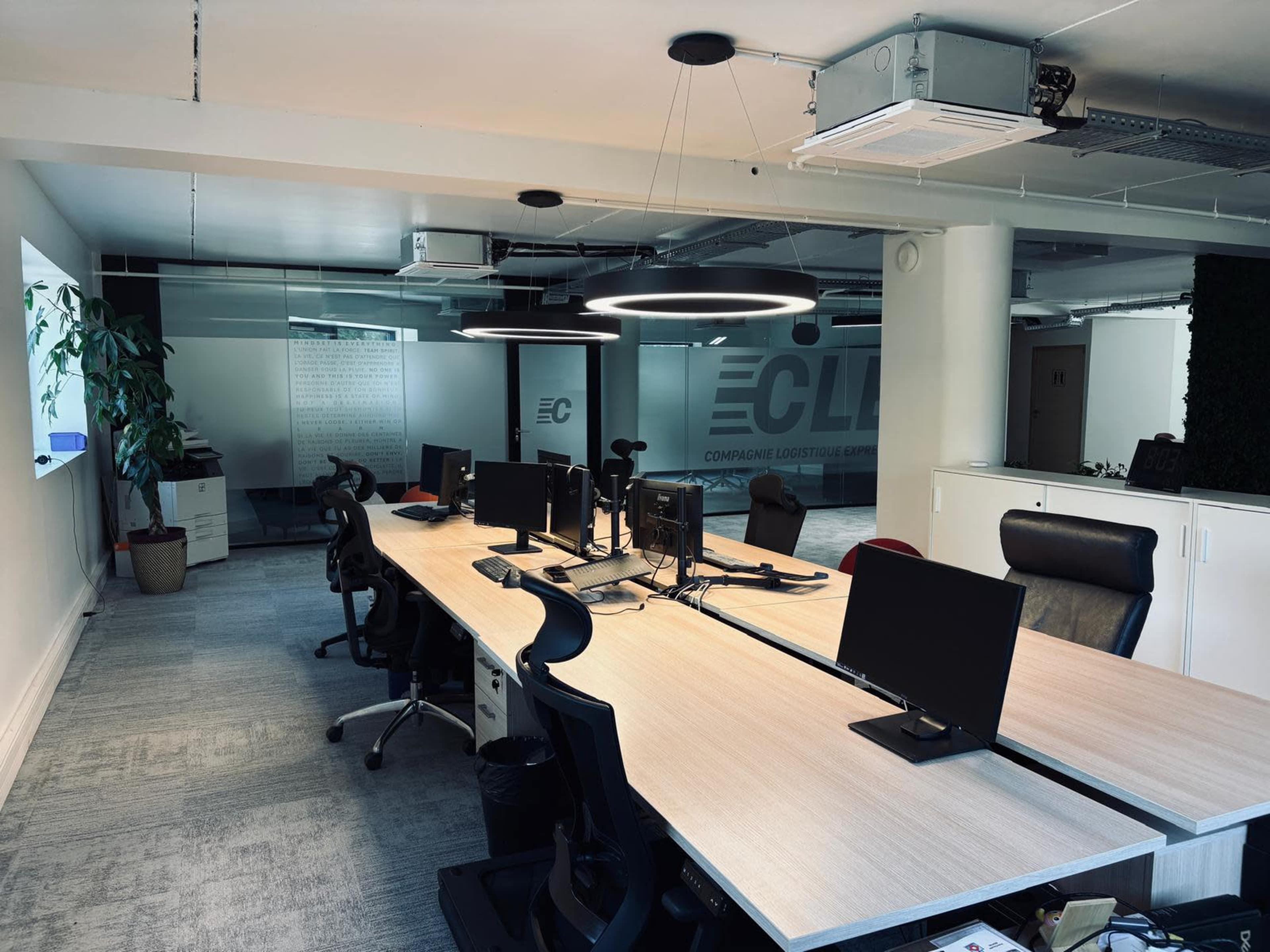 The image shows a modern office space with multiple desks, each equipped with computer monitors, along with a plant and glass partition featuring the company logo.