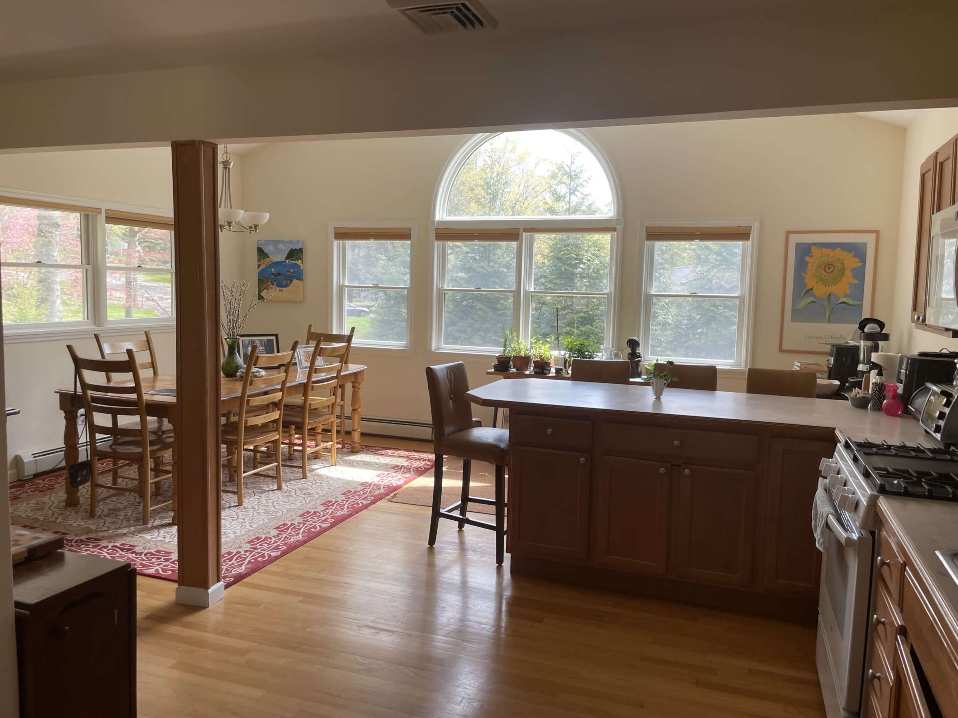 A bright kitchen with wooden cabinets opens to a dining area featuring a large window and a table set for six.