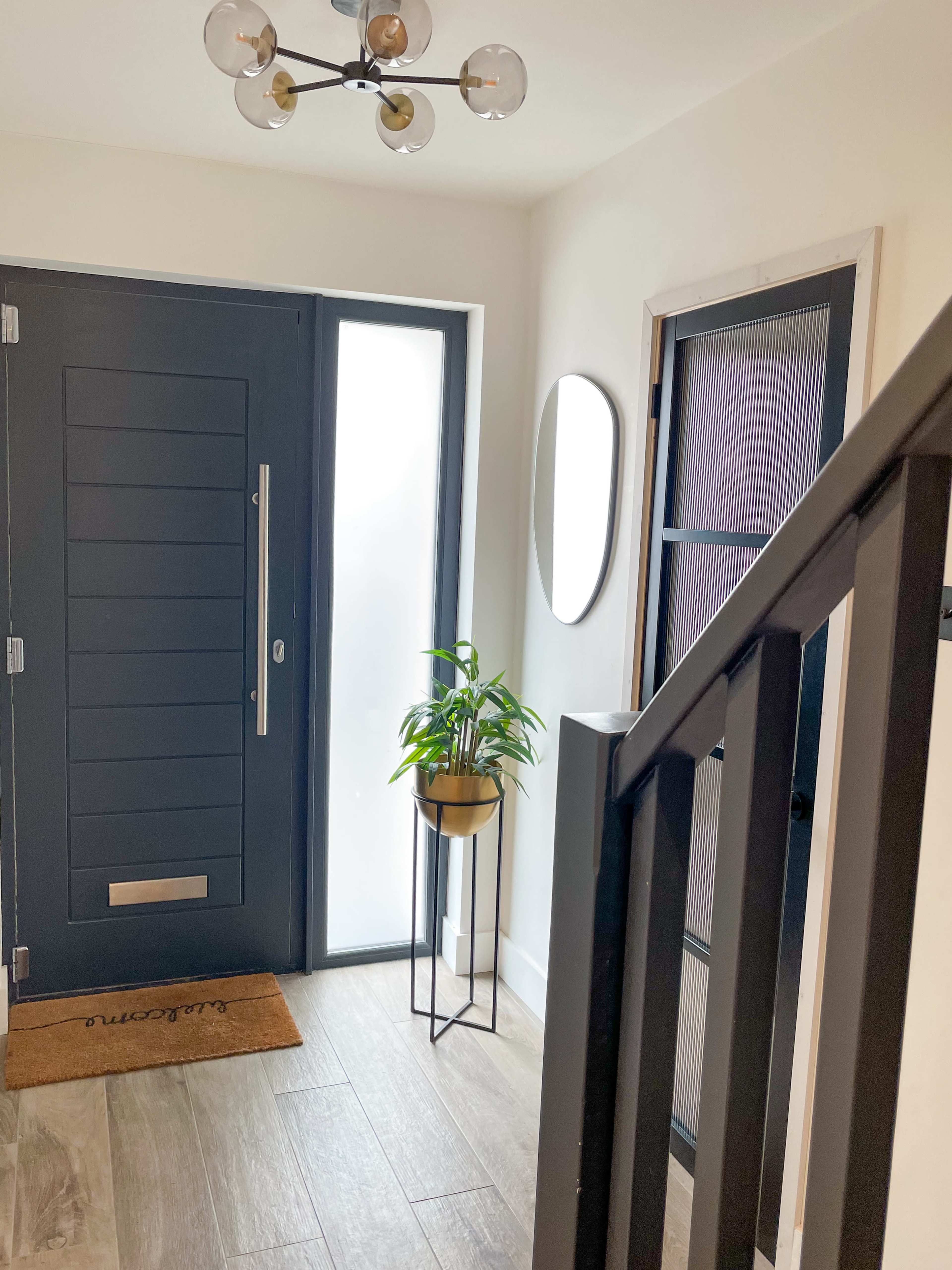 A hallway features a dark front door, a round mirror, a potted plant on a stand, and a staircase leading upwards.