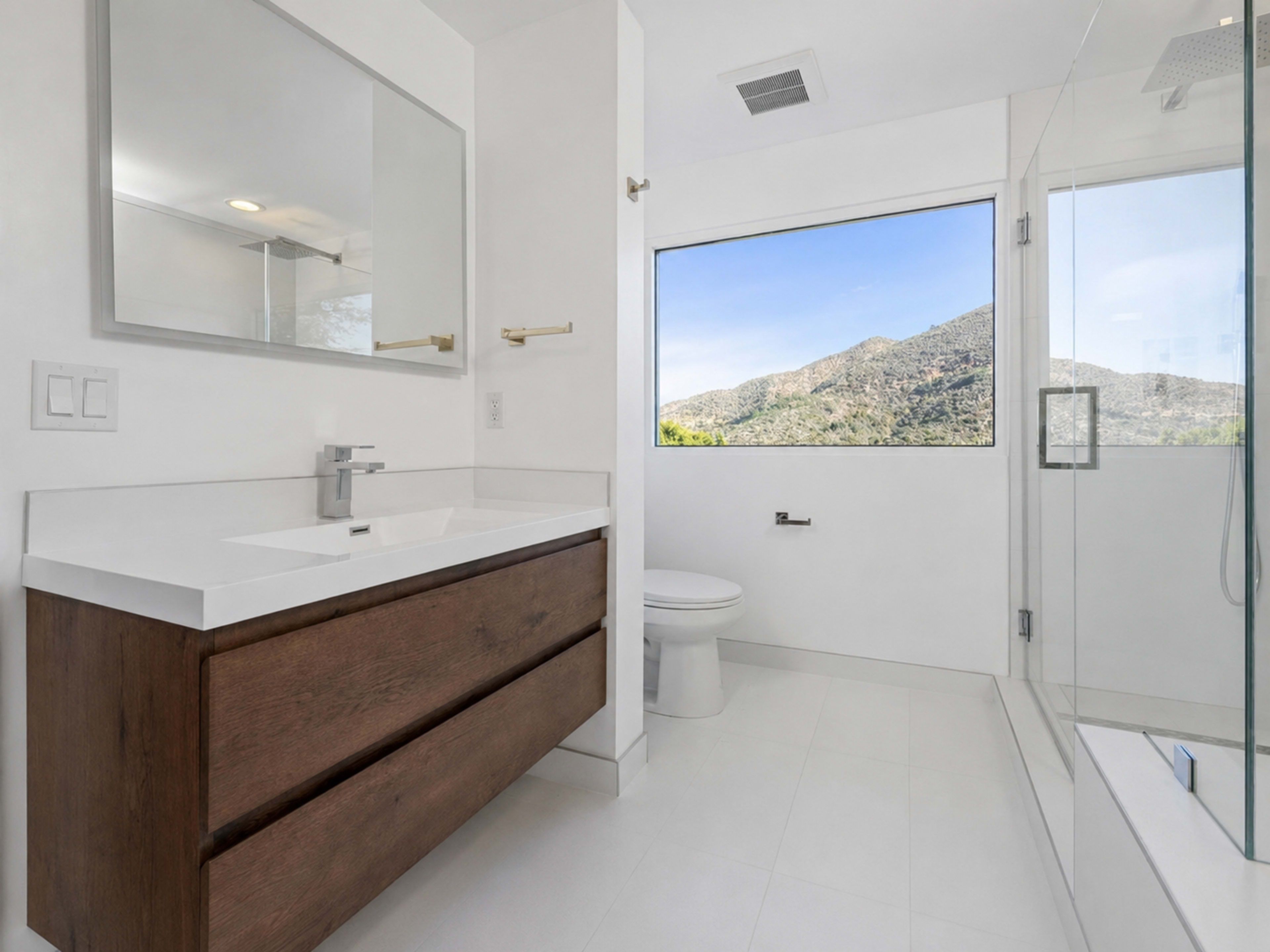 The image shows a modern bathroom featuring a white vanity with a wooden base, a toilet, and a glass-enclosed shower, all with a large window offering a view of a mountain landscape.