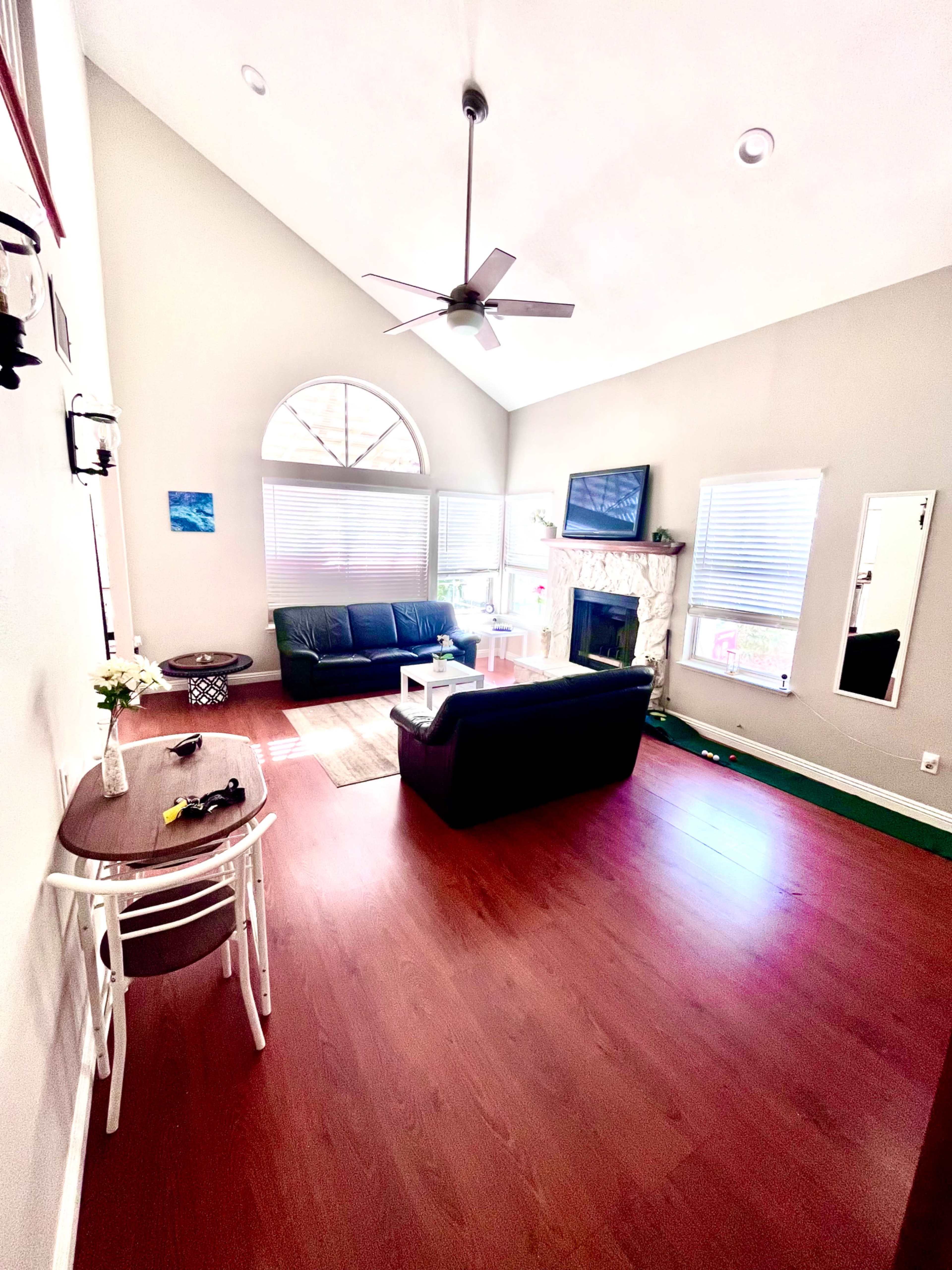 The room features a high ceiling, a black leather sofa, a fireplace with a TV above it, and a small table near a large window.