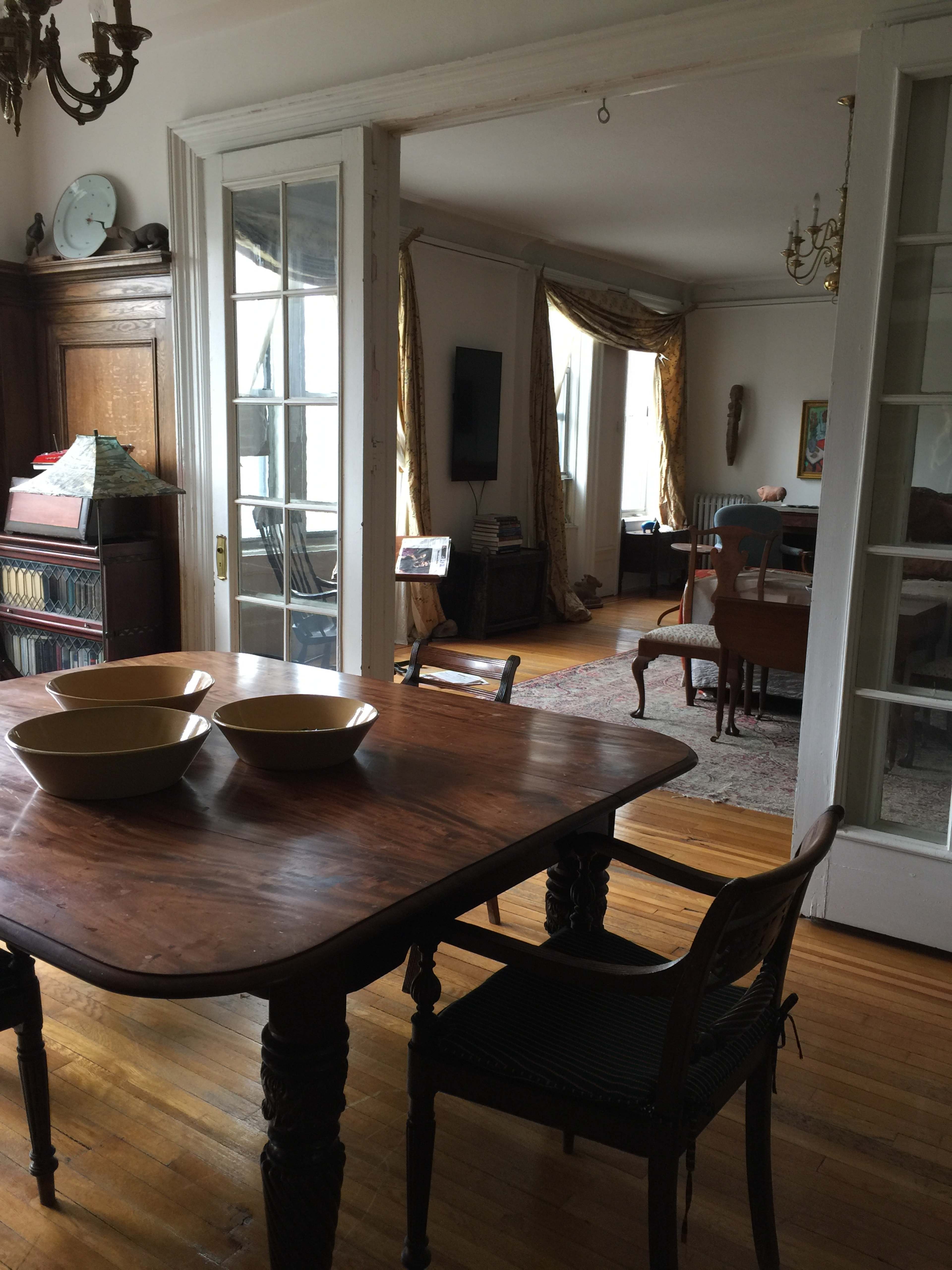 A wooden table with two bowls sits in a dining area, leading to a living room visible through glass-paned doors.