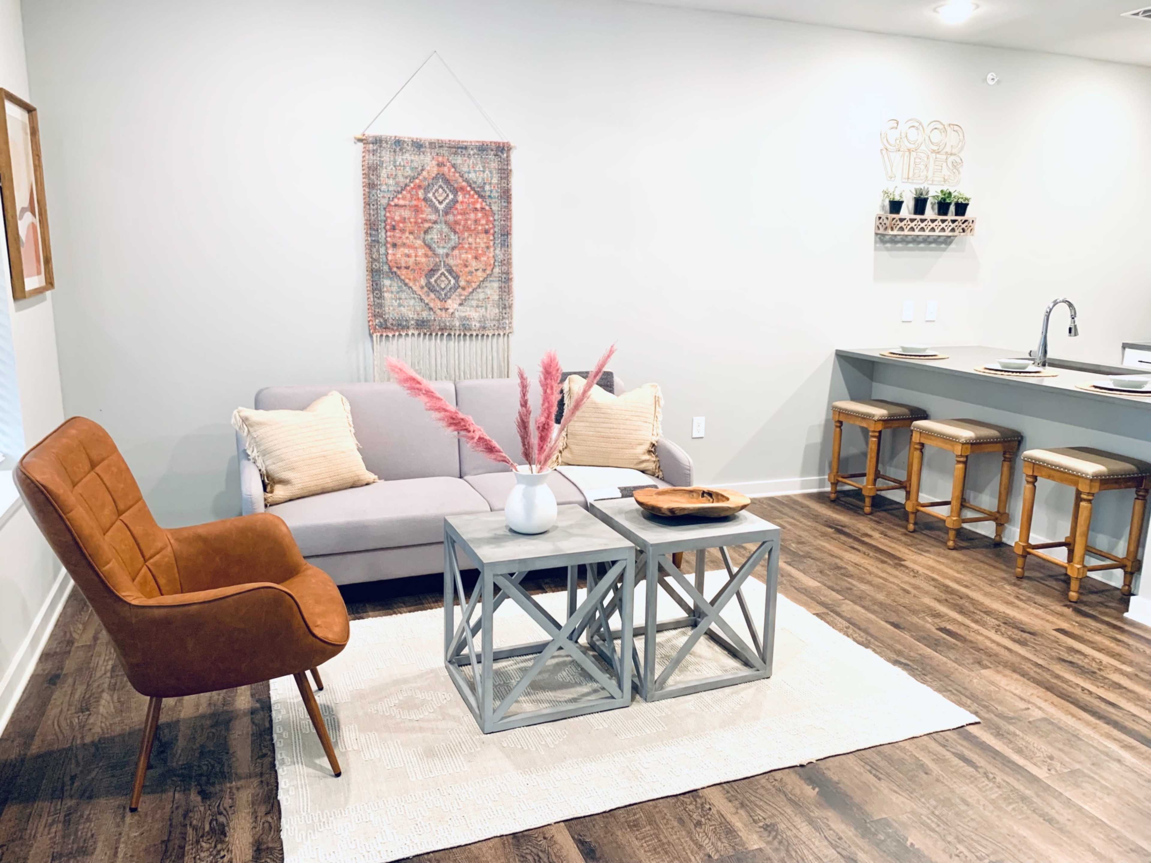 A modern living room with a gray sofa, a wooden coffee table, a decorative wall hanging, and a kitchen bar in the background.