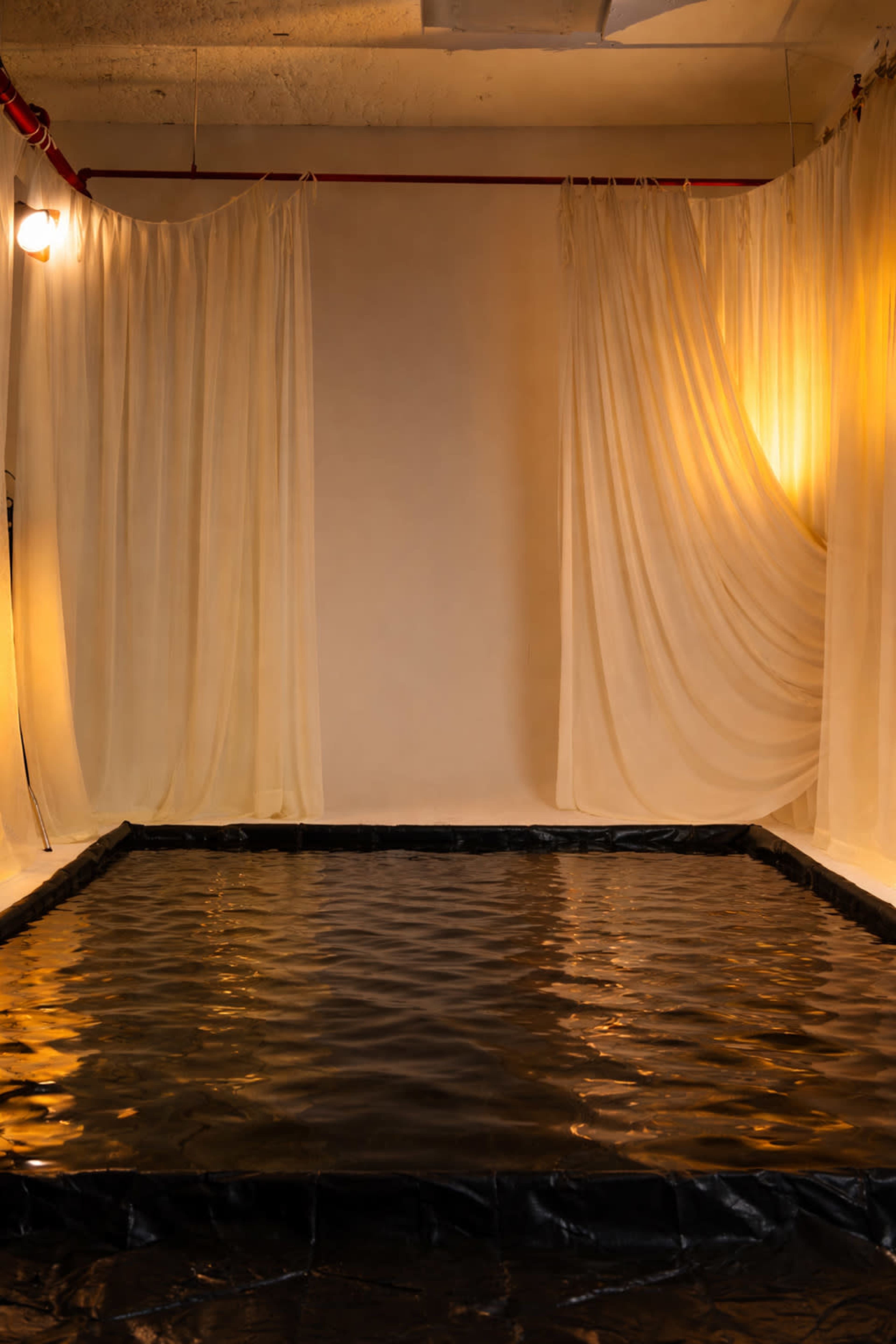 The image shows a minimal indoor space featuring a shallow pool with a black tarp lining, surrounded by flowing white curtains.