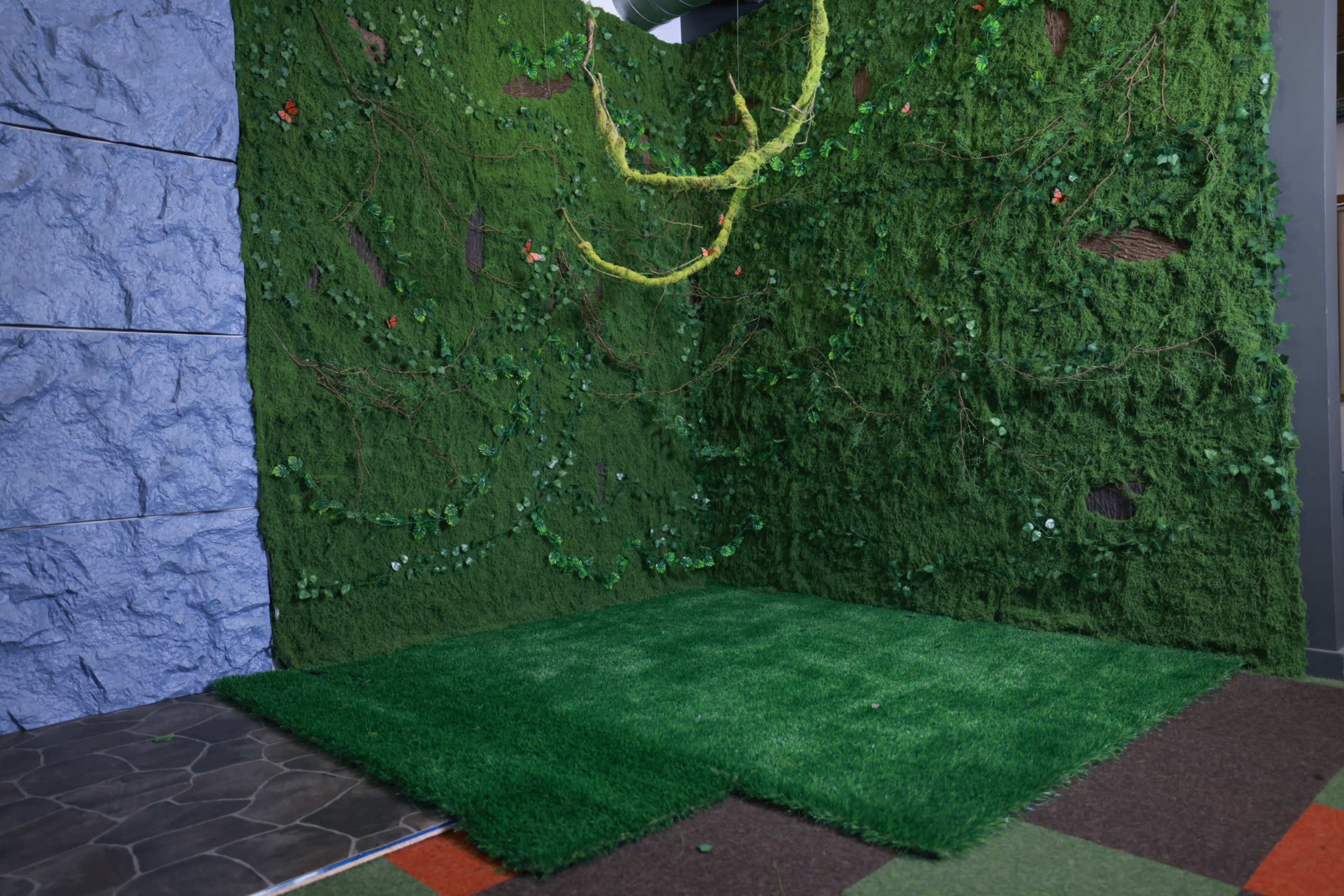 The image shows a corner of a room decorated with artificial green foliage and grass, creating a nature-themed backdrop against textured walls.