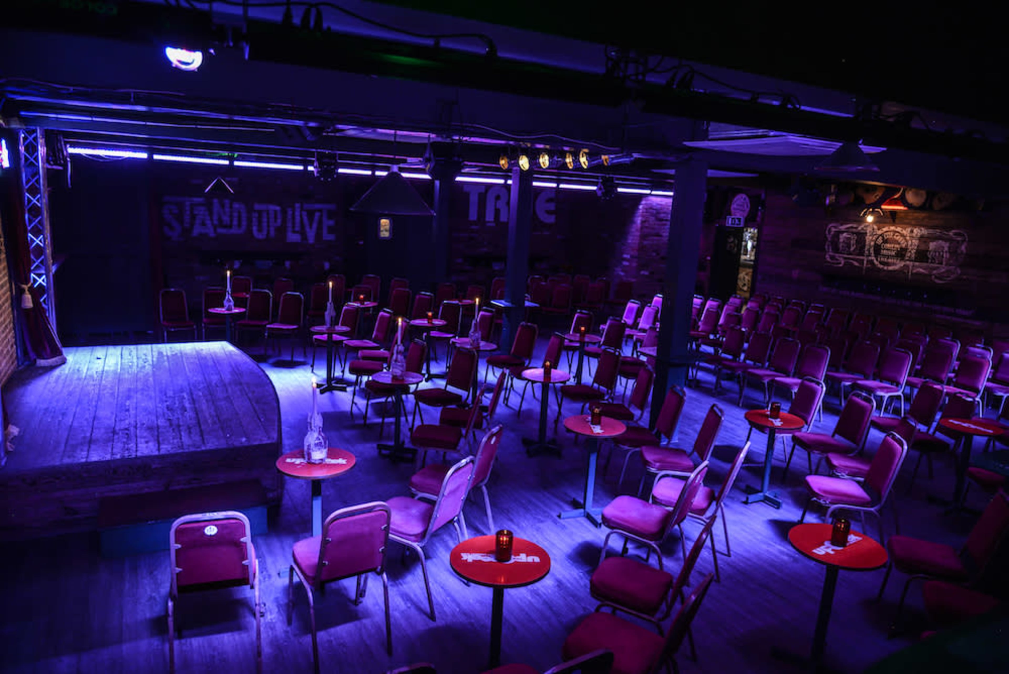 The image shows an empty comedy club with red circular tables and chairs, illuminated by purple lighting, featuring a small stage at one end.