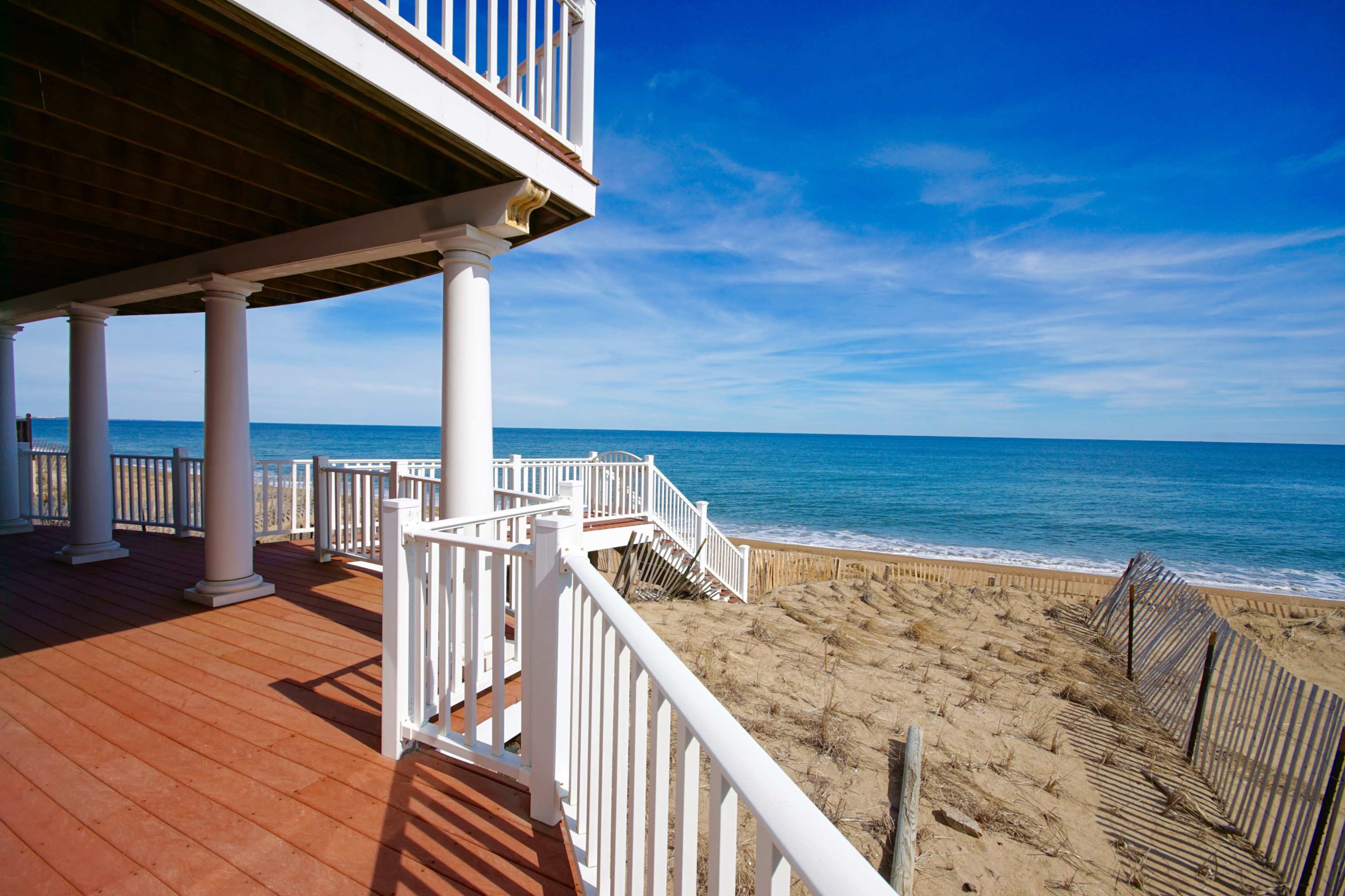 A wooden deck with white railings overlooks a sandy beach and the ocean under a clear blue sky.