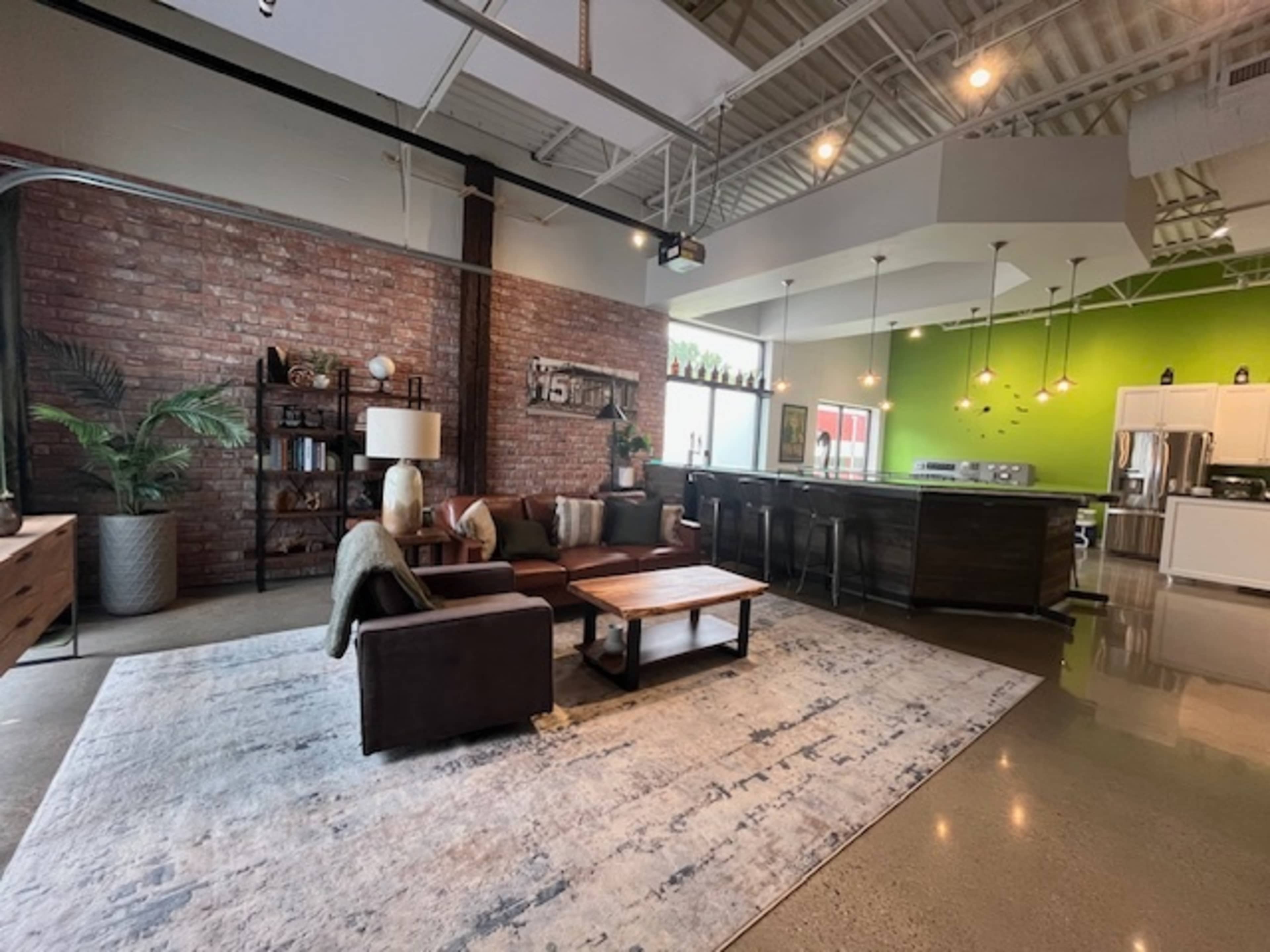 The image shows a modern living area with a brown sofa, a wooden coffee table, and a large area rug, adjacent to a kitchen featuring green walls and stainless-steel appliances.