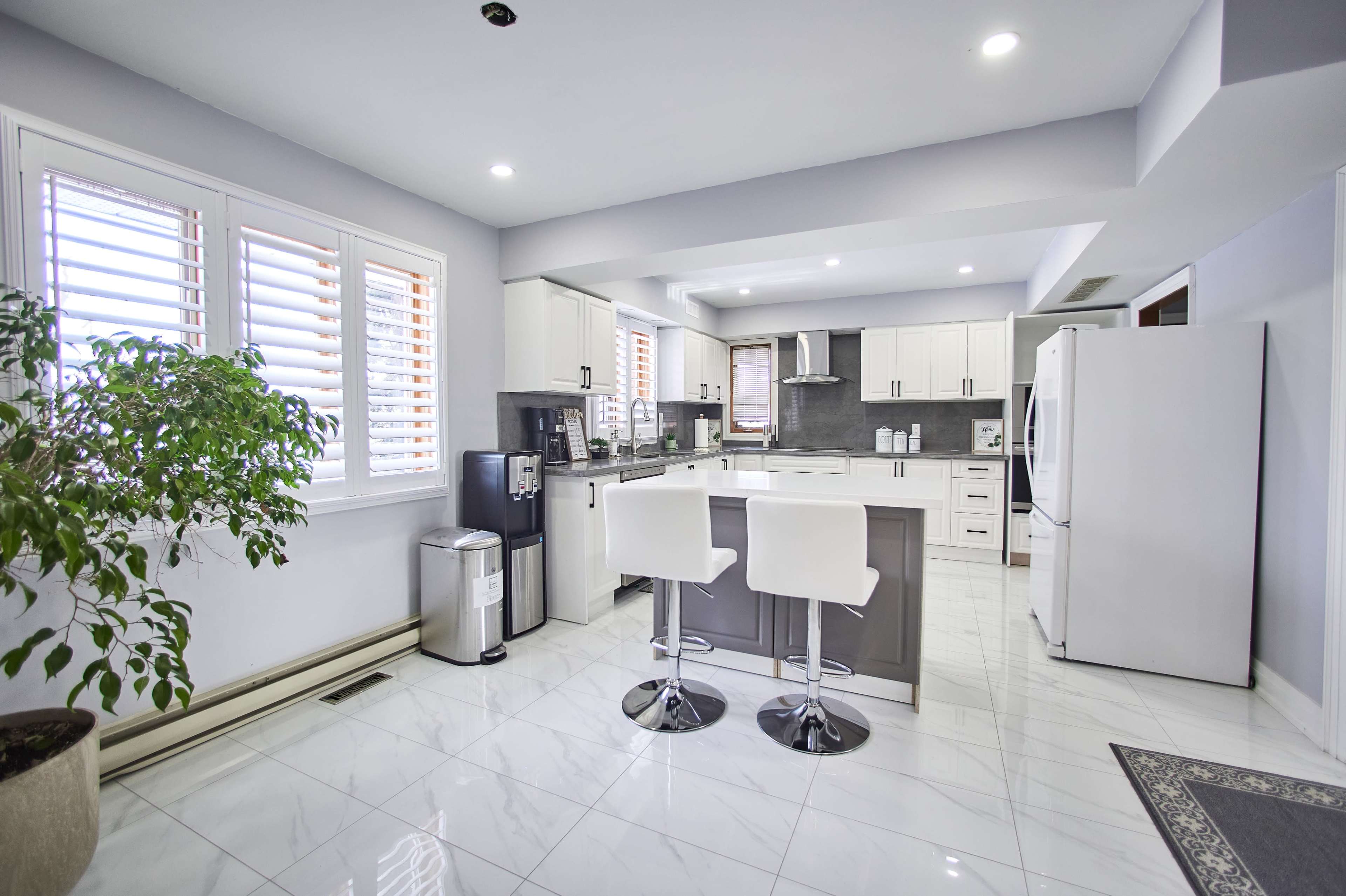 A modern kitchen featuring white cabinets, a gray island with bar stools, and large windows with shutter blinds.