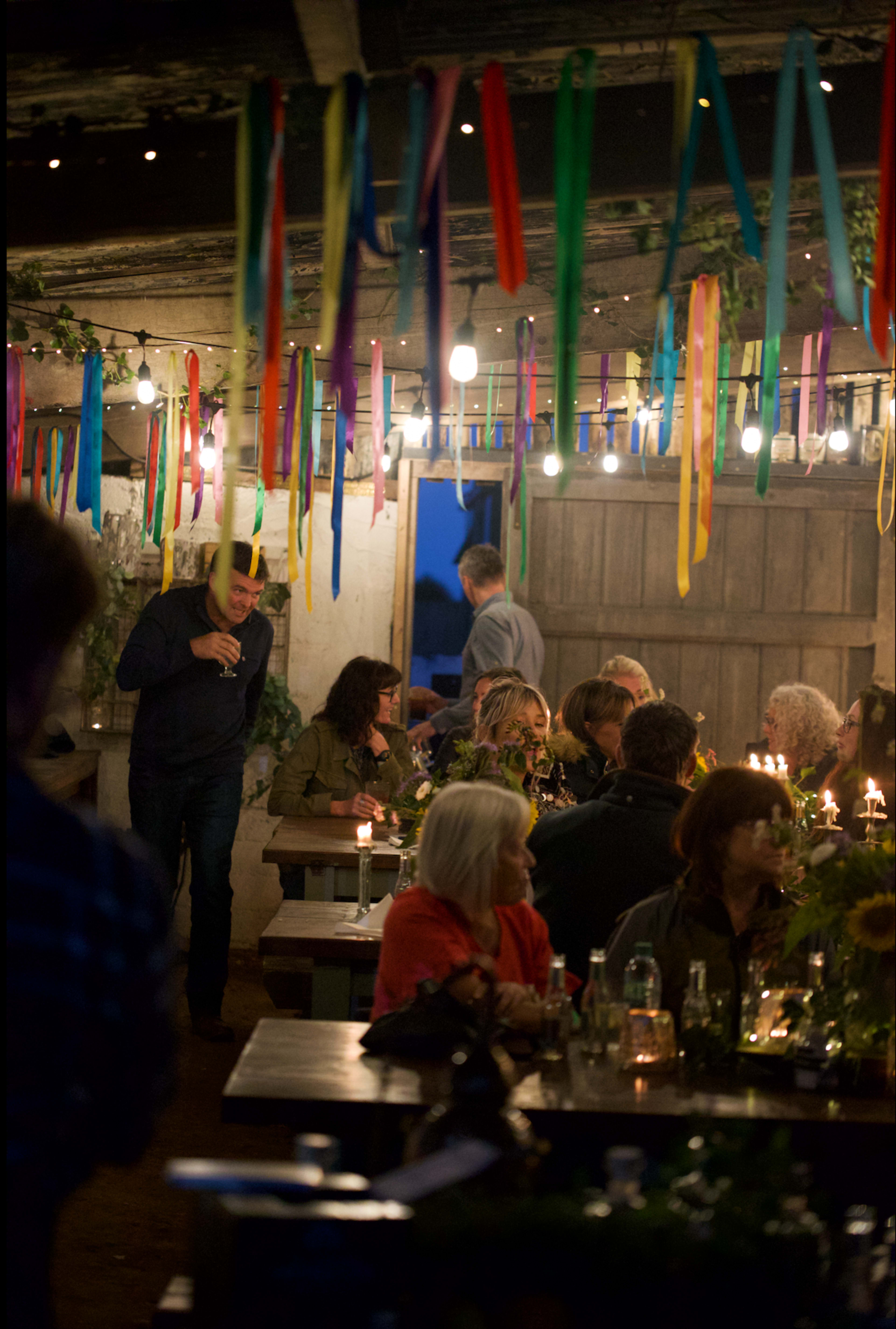 A group of people gather around tables decorated with candles and sunflowers in a dimly lit room adorned with colorful ribbons.