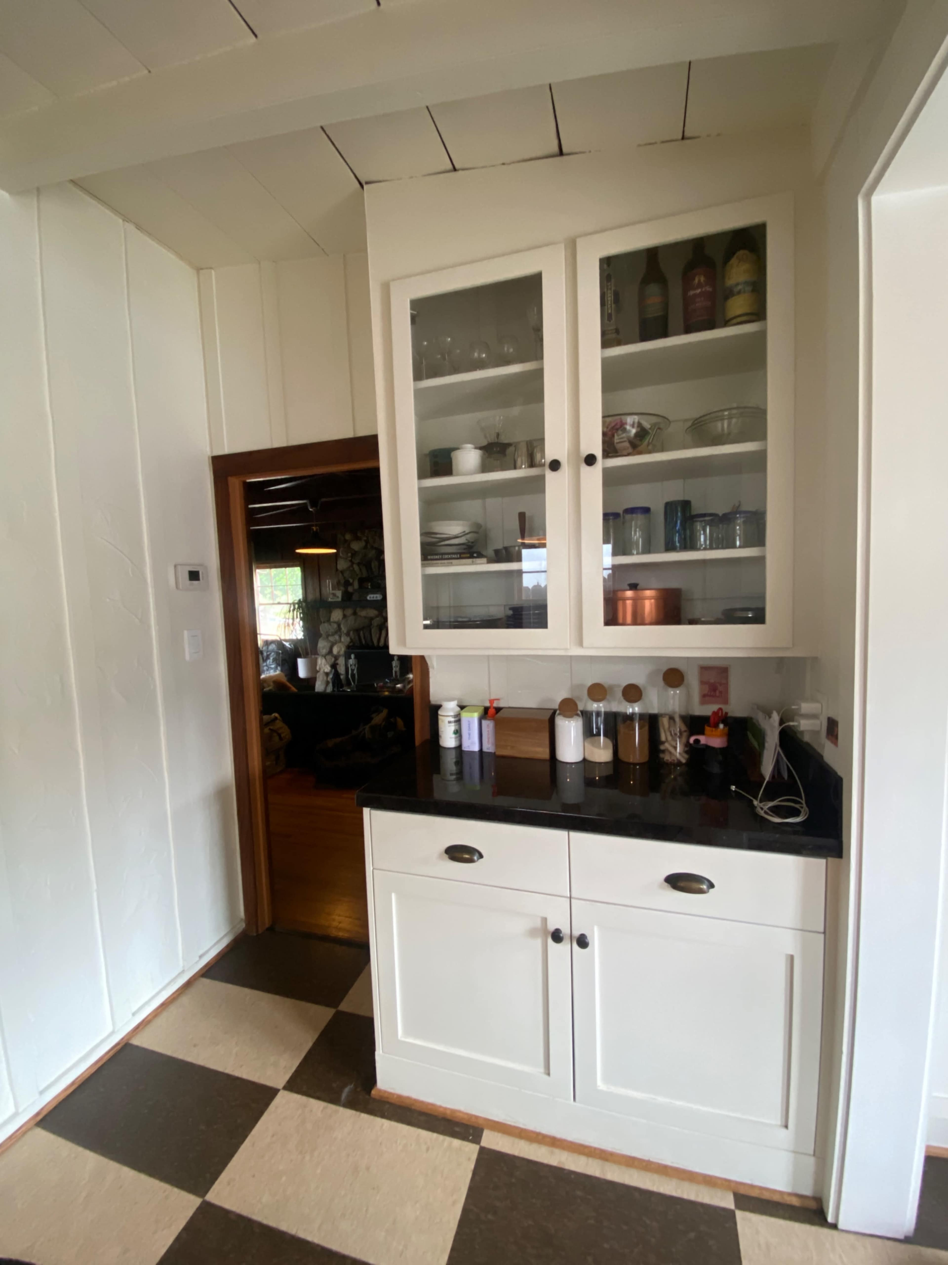 The image shows a kitchen corner with a cabinet featuring glass doors displaying various dishes and glassware, adjacent to a doorway leading to another room.