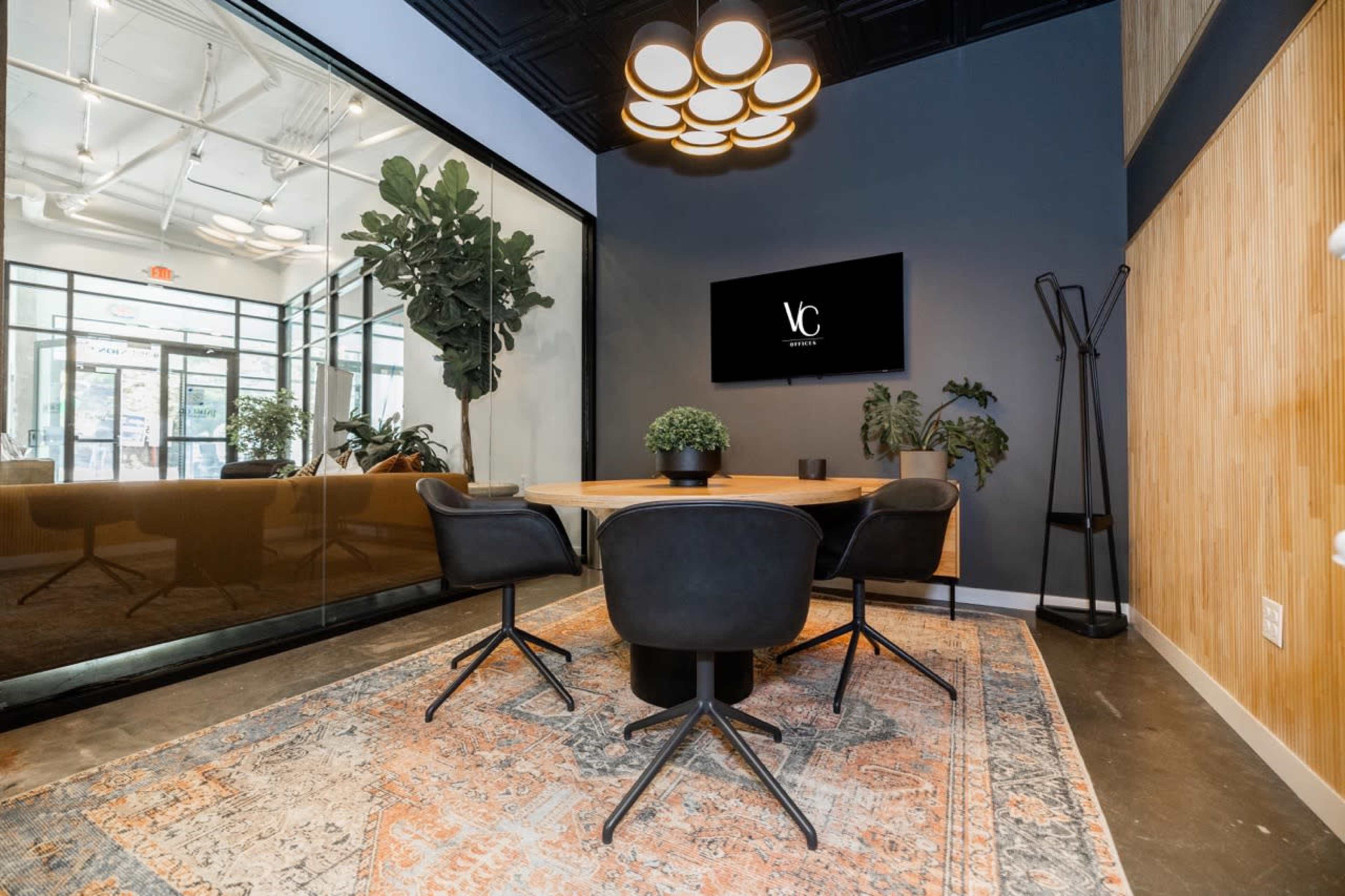 A modern conference room features a wooden table surrounded by black chairs, with a flat-screen TV on the wall and large windows allowing natural light to enter.