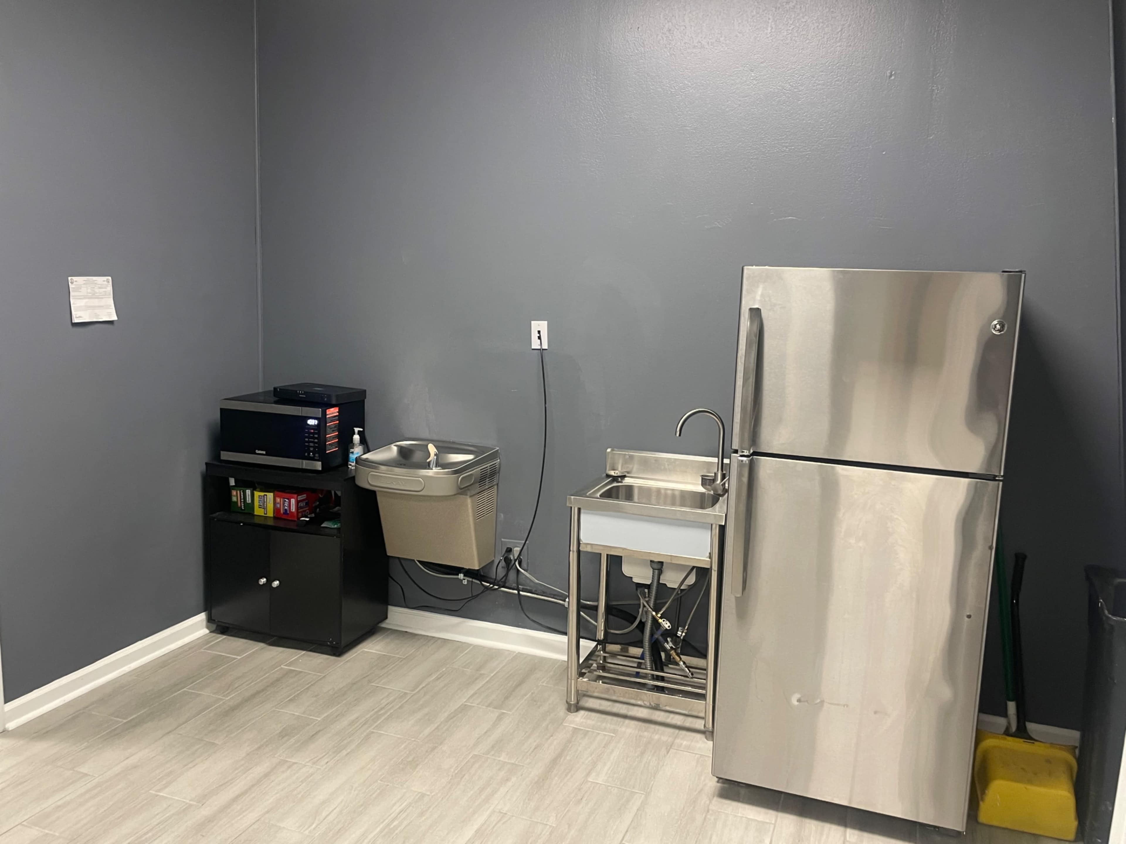 A simple kitchen area with a stainless steel refrigerator, a sink with a metal countertop, a microwave, and a black cabinet against a gray wall.