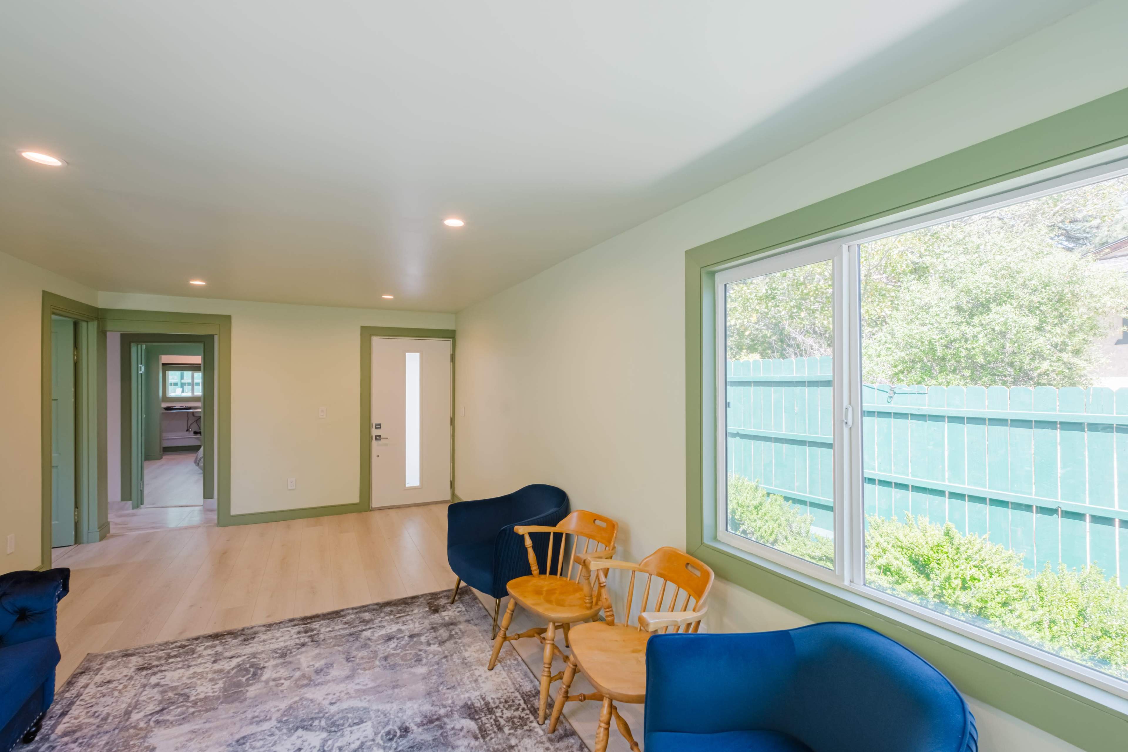 The image shows a light-colored living space with two blue chairs, two wooden stools, a window overlooking a green fence, and doorways leading to other rooms.