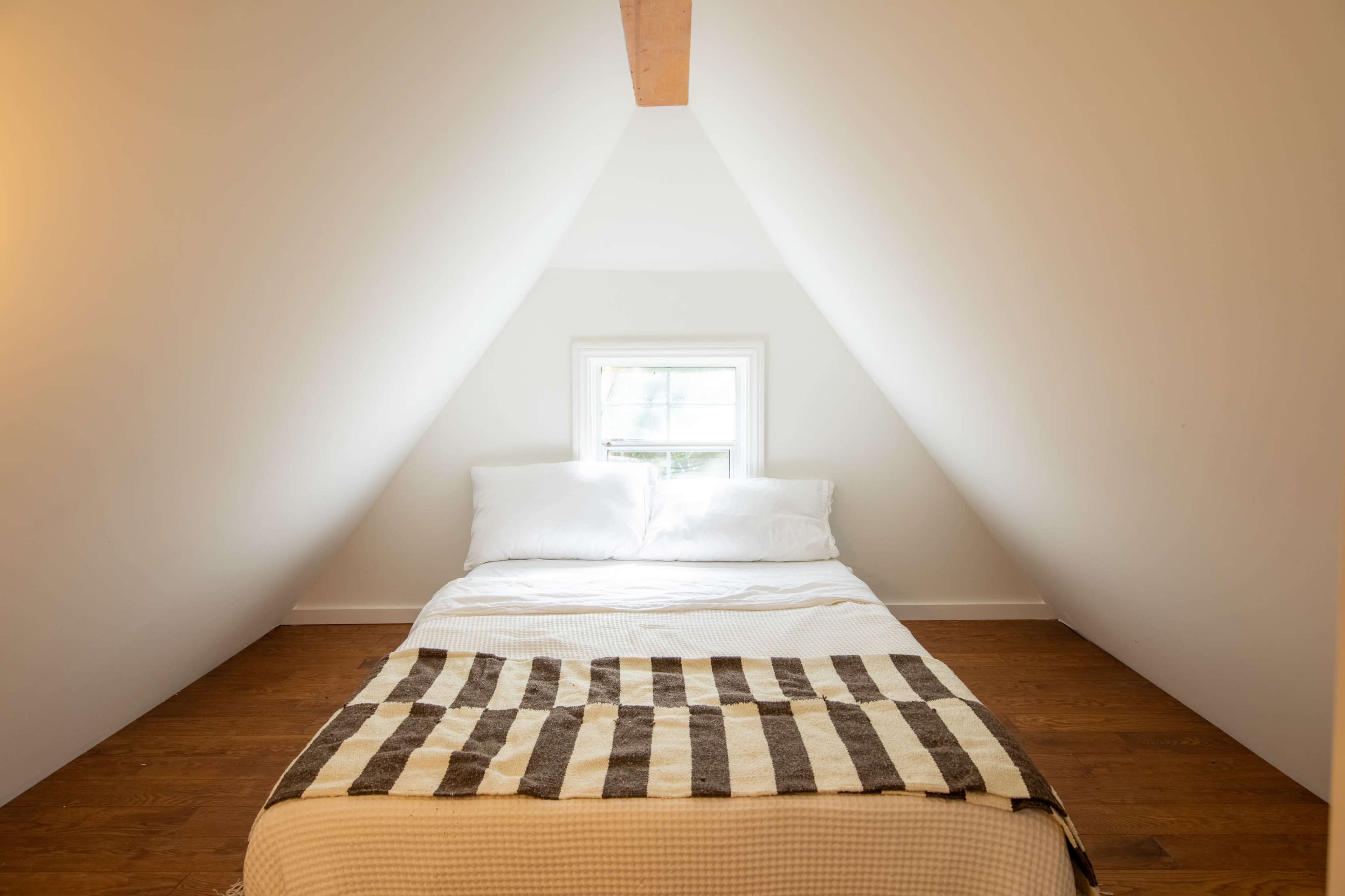 A bed with white bedding and a patterned throw is positioned against a sloped wall under a small window in an attic room.