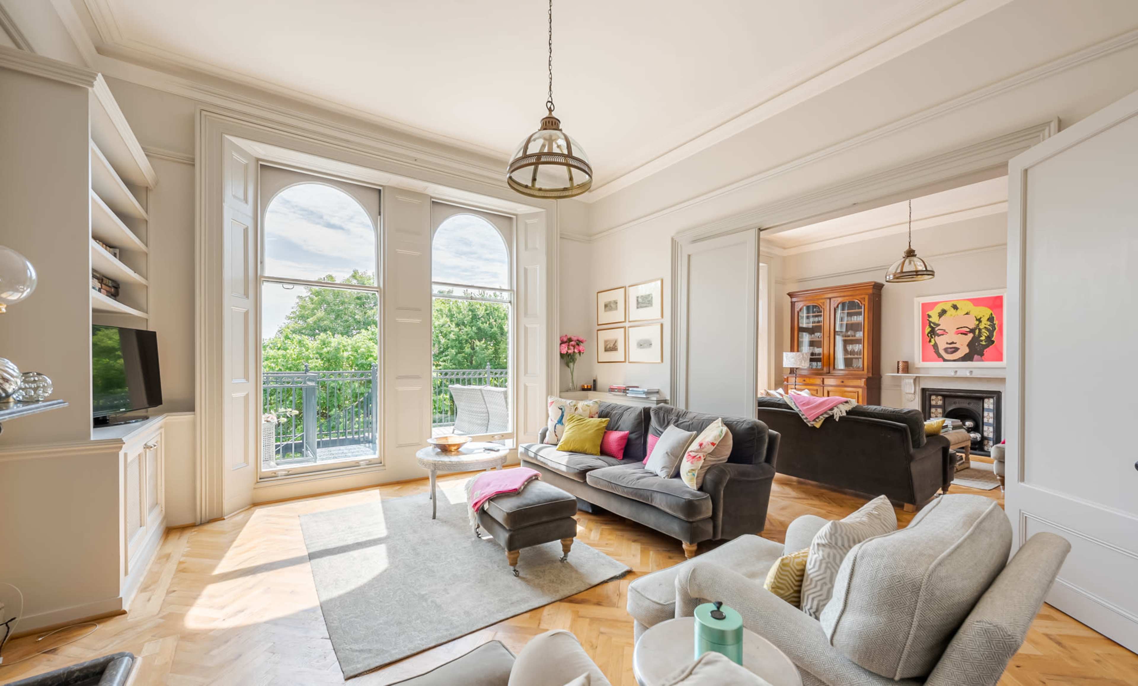 The image shows a bright living room featuring large windows, a gray sofa, a coffee table, and a view of greenery outside.