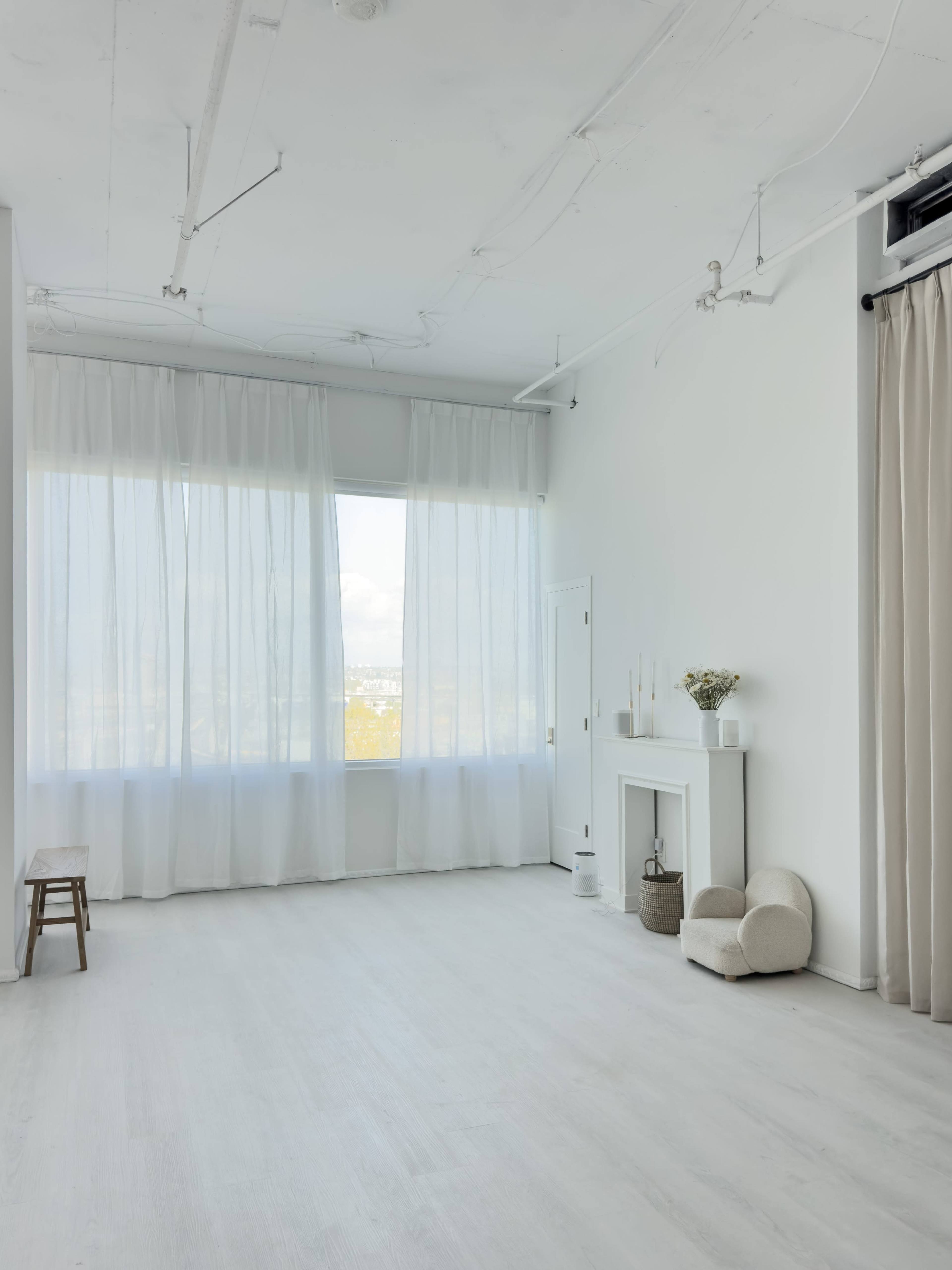 The image shows a bright, minimalist room with large windows draped in sheer white curtains and light-colored wooden flooring.