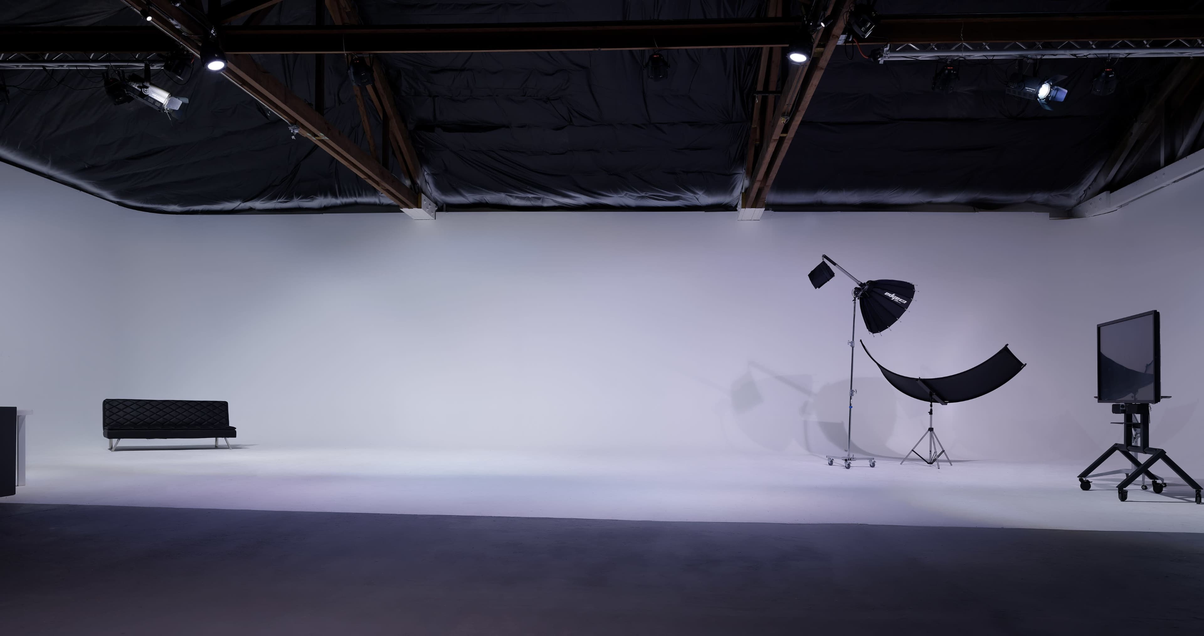The image shows a spacious studio featuring a black couch on one side, various lighting equipment, and a plain white wall.