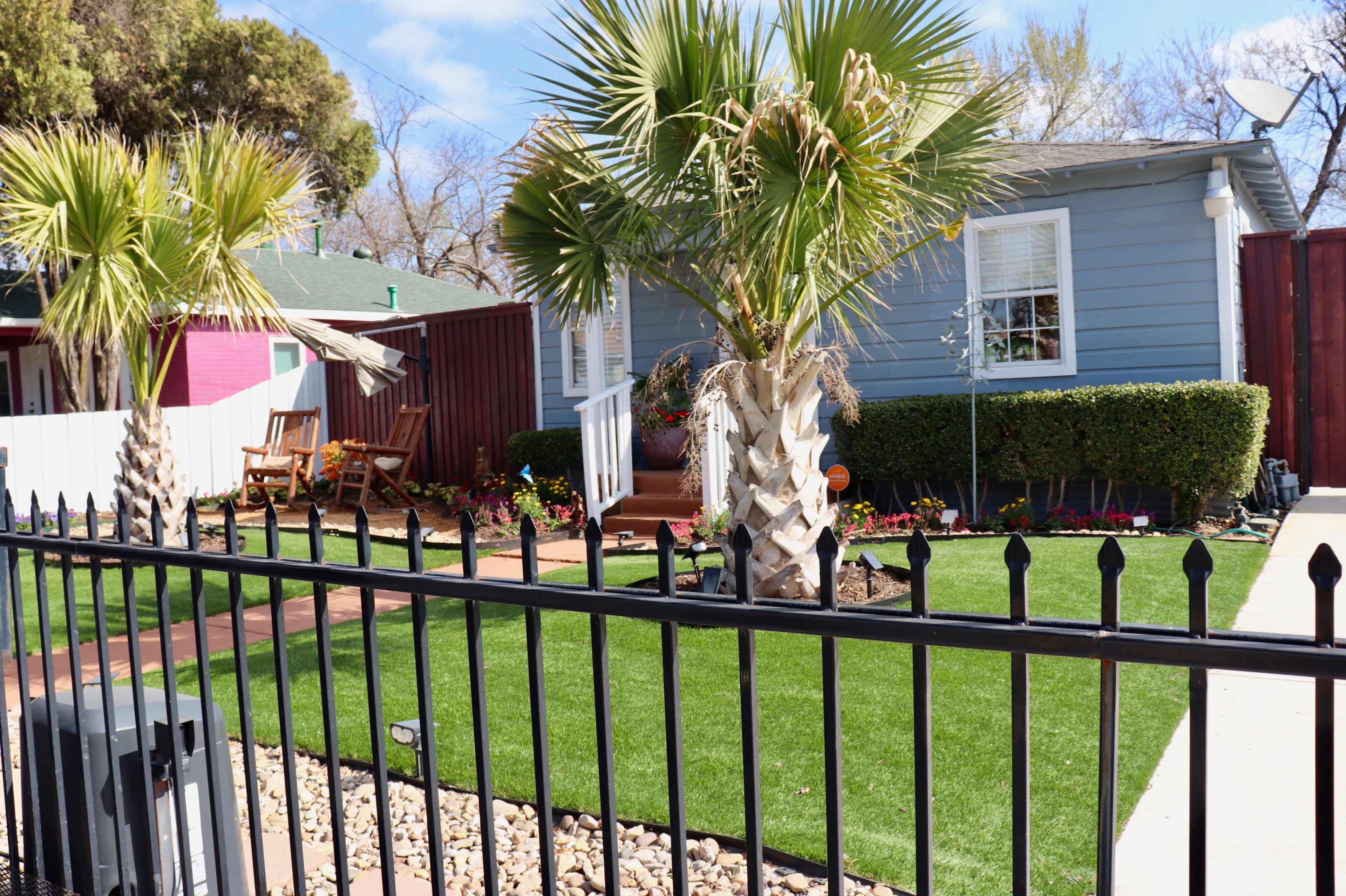 A house with a light blue exterior, surrounded by a well-maintained yard featuring palm trees, a green lawn, and a decorative fence.