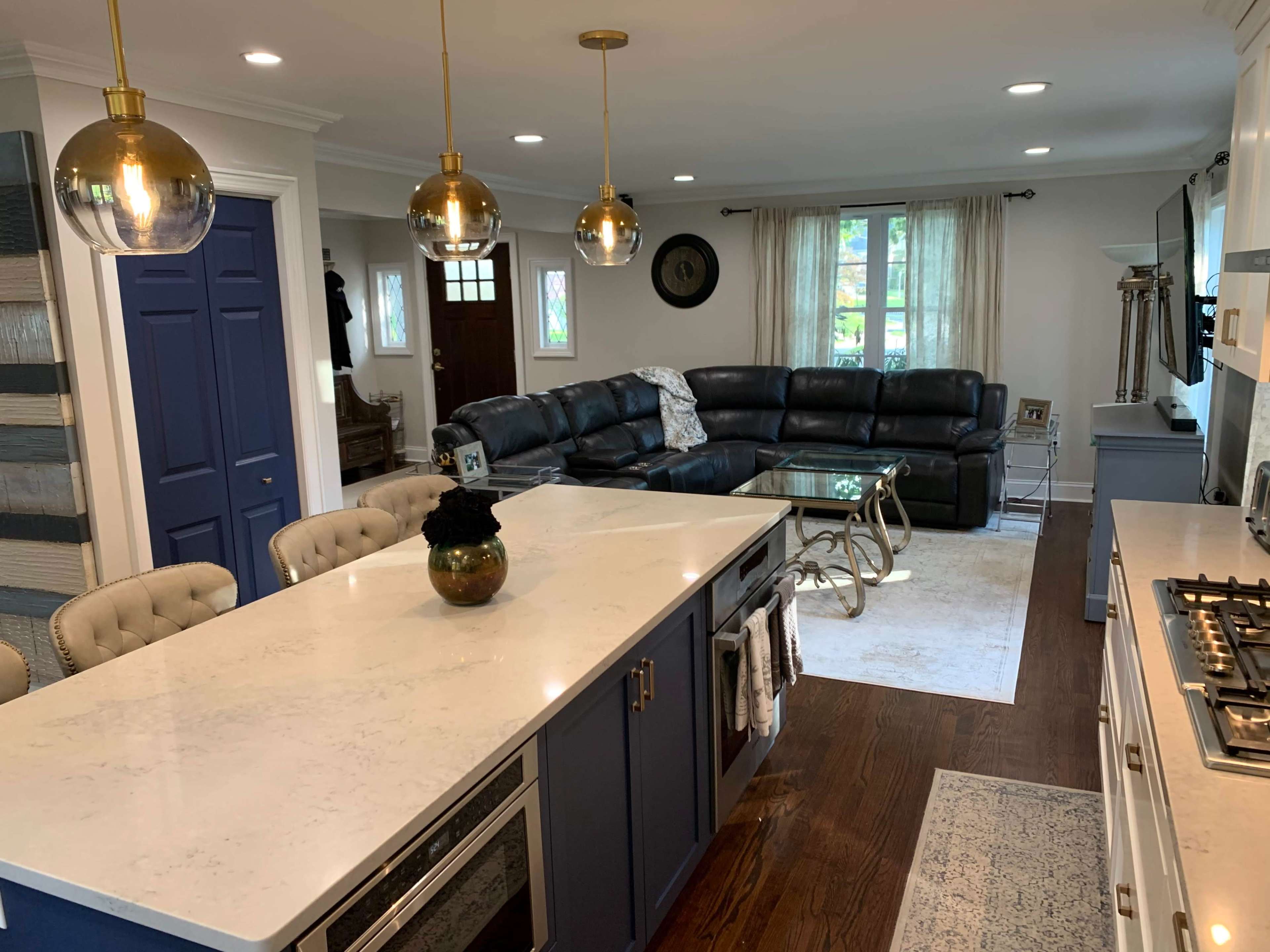 A modern kitchen with a marble countertop island, a black leather sectional sofa, and pendant lights illuminating the space.