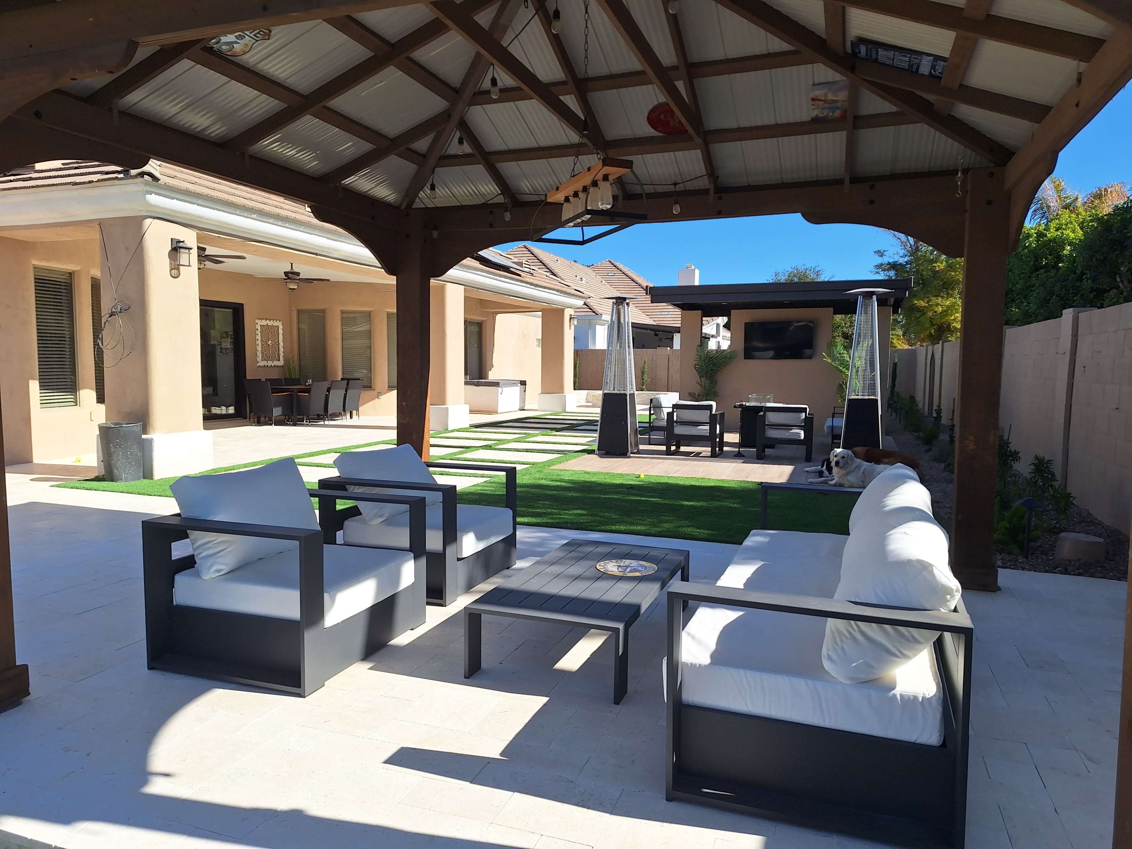The image shows a covered patio area with modern outdoor furniture overlooking a well-maintained backyard featuring grass and a fire pit.