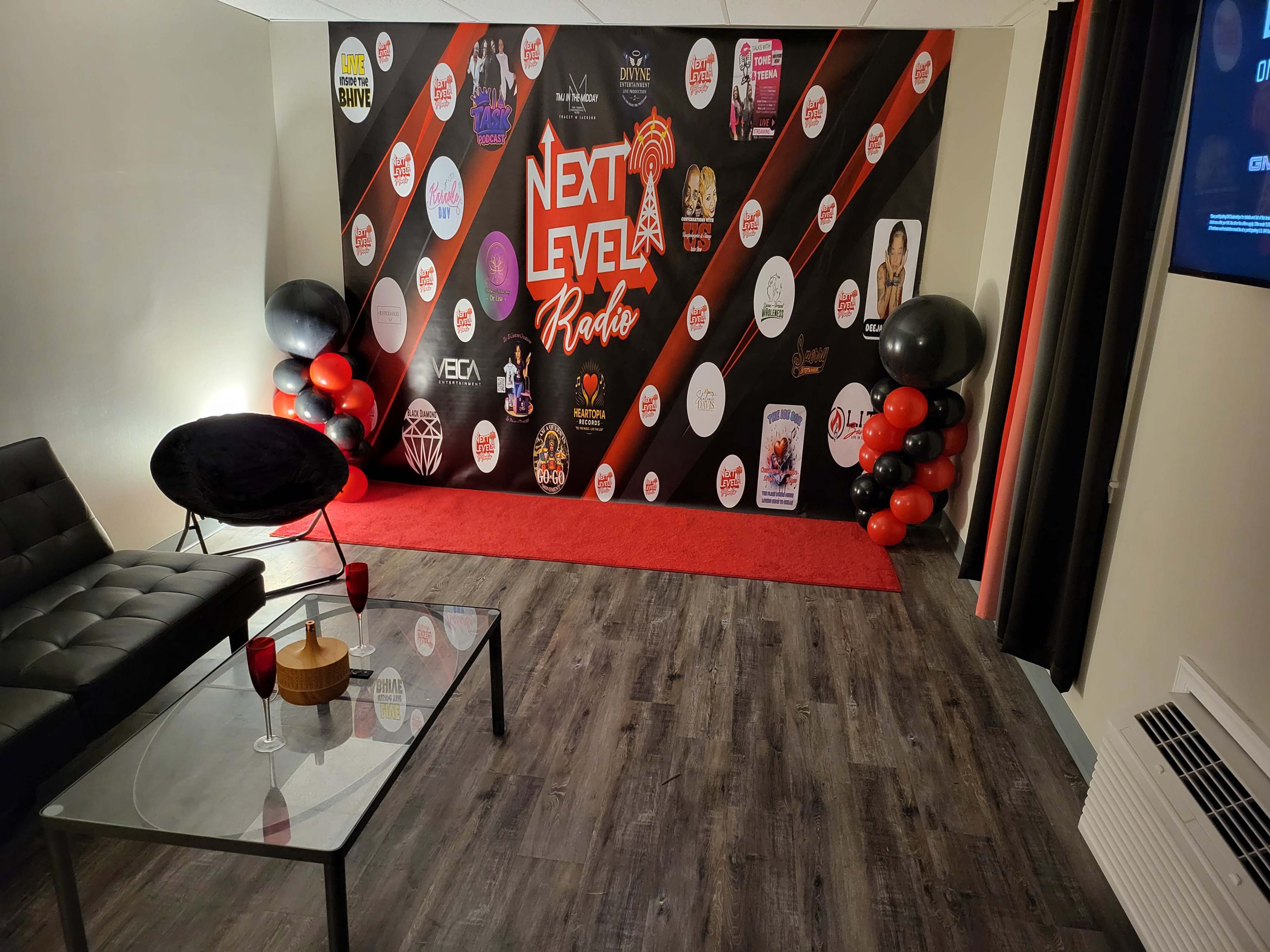 The image shows a decorated room with a black and red backdrop featuring various circular logos, a red carpet, a black couch, a glass coffee table, and decorative balloons on one side.