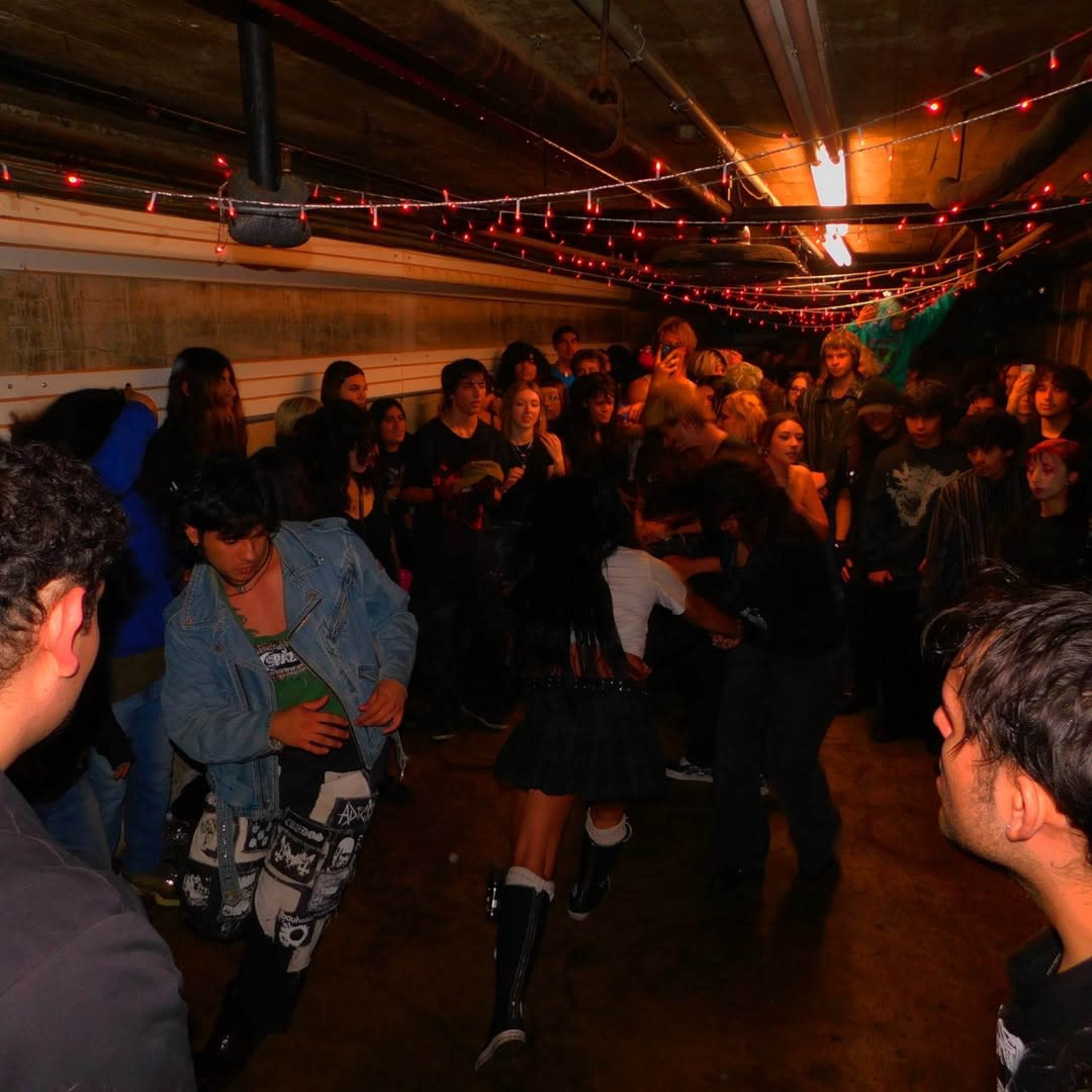 A large group of young people gathers and participates in a lively event in a dimly lit hallway adorned with string lights.