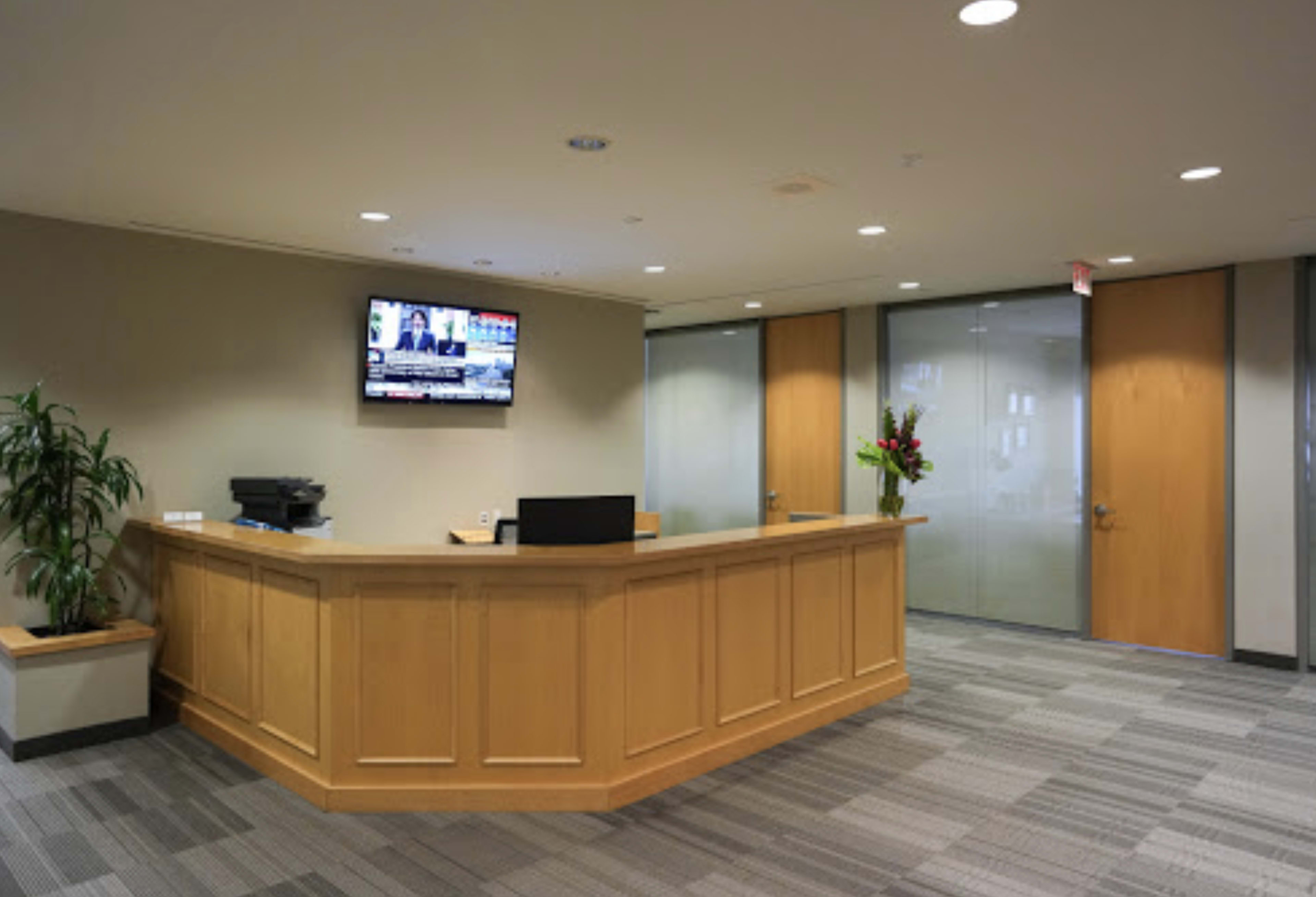 A modern reception area features a wooden front desk, a wall-mounted television, and glass doors leading to another room.