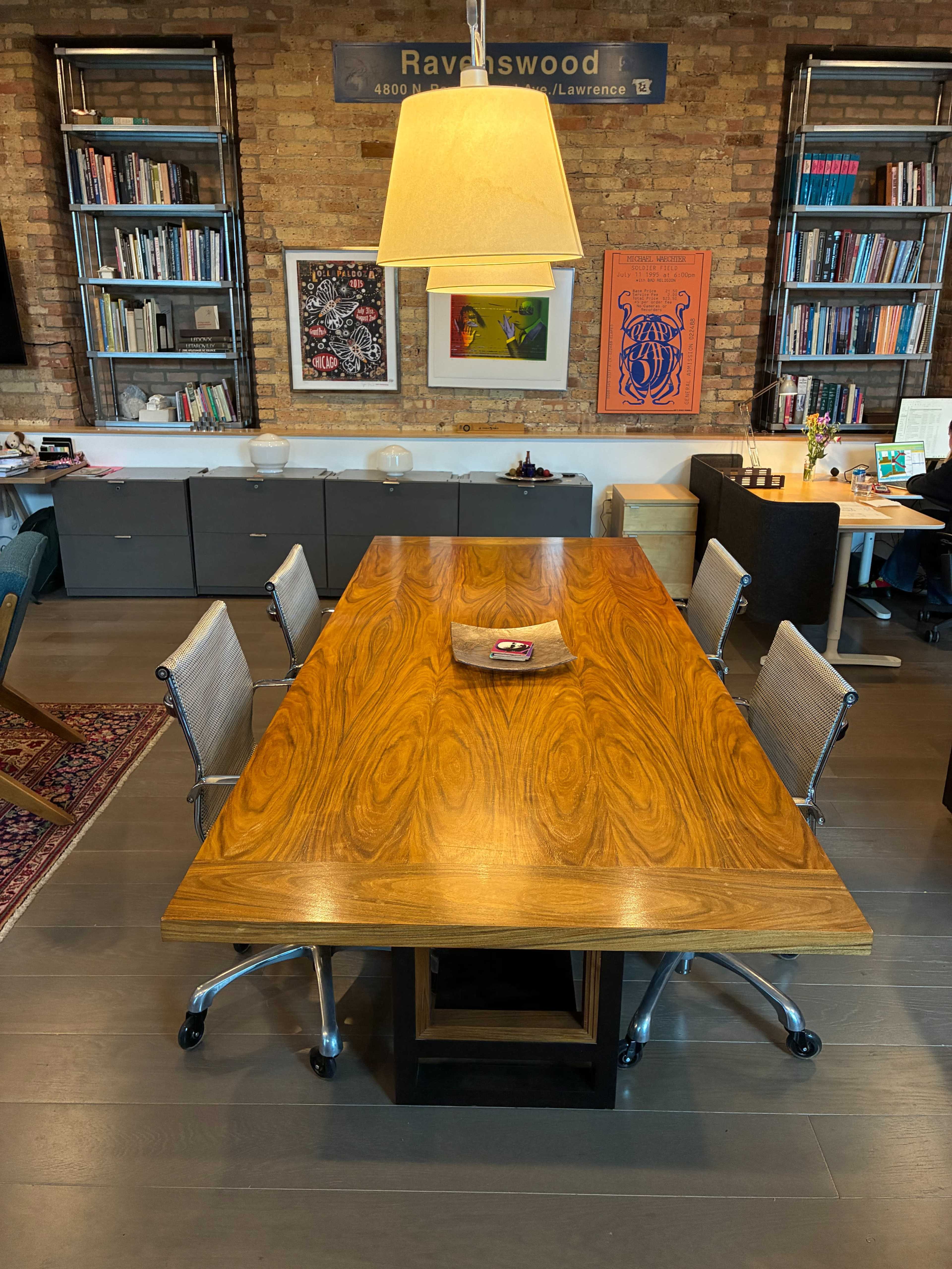 A large wooden conference table is surrounded by four metal and mesh chairs in a well-lit office space with brick walls and shelves filled with books and artwork.