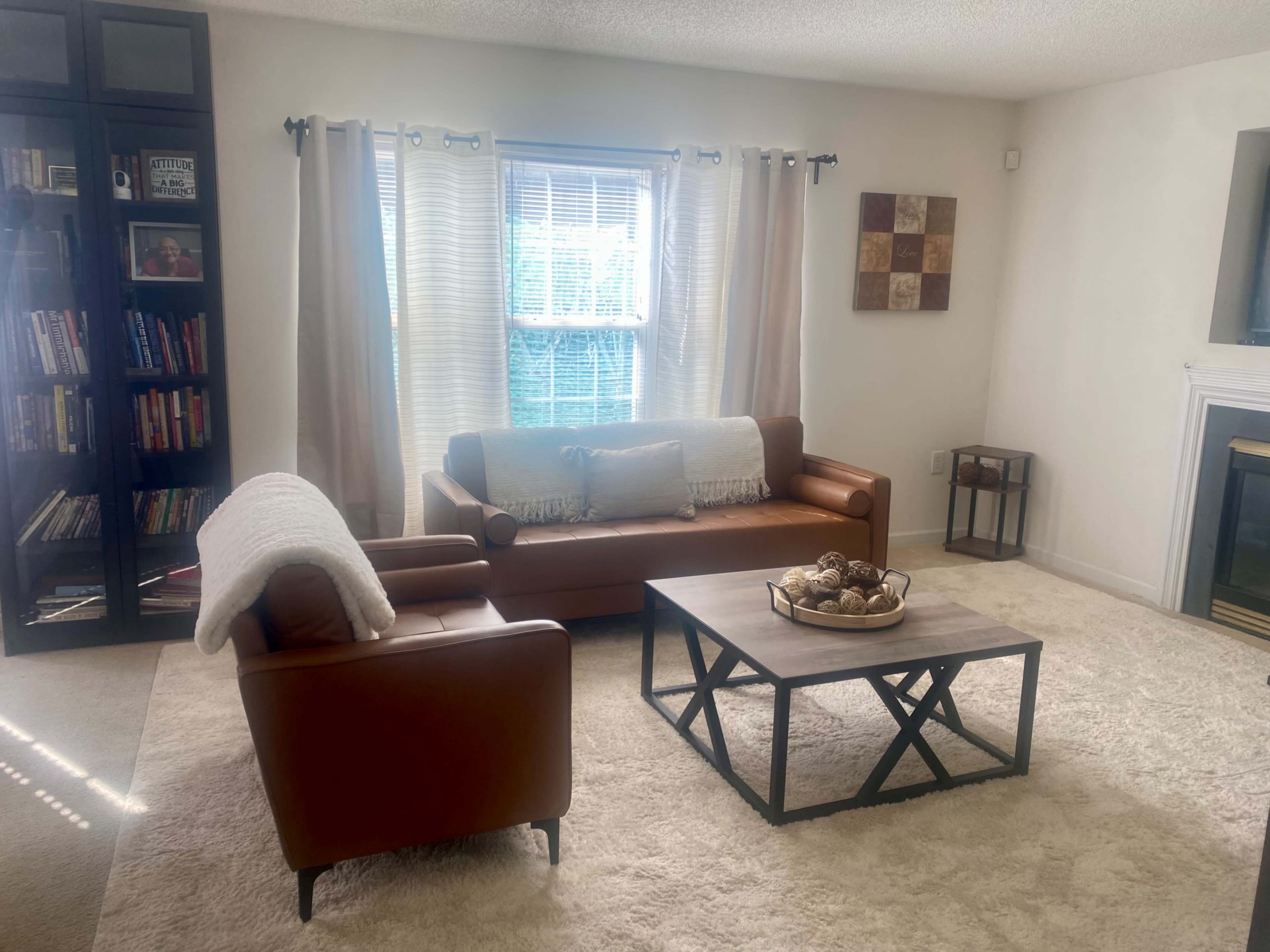 The living room features a brown leather sofa and an armchair, a wooden coffee table with decorative items, a bookshelf filled with books, and large windows with sheer curtains.