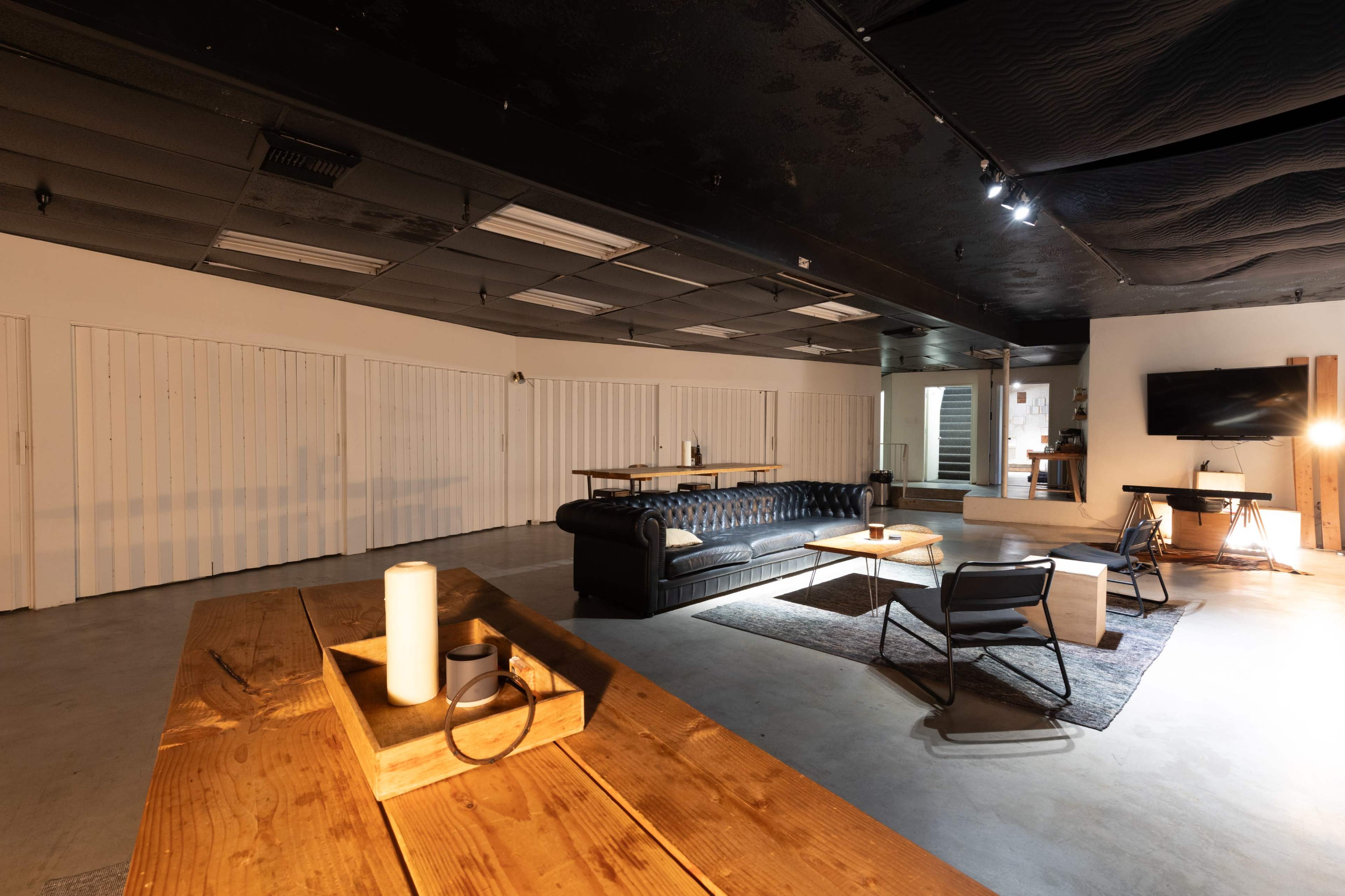 The image shows a modern interior space with a dark ceiling, wooden tables, a black leather sofa, and various seating arrangements.