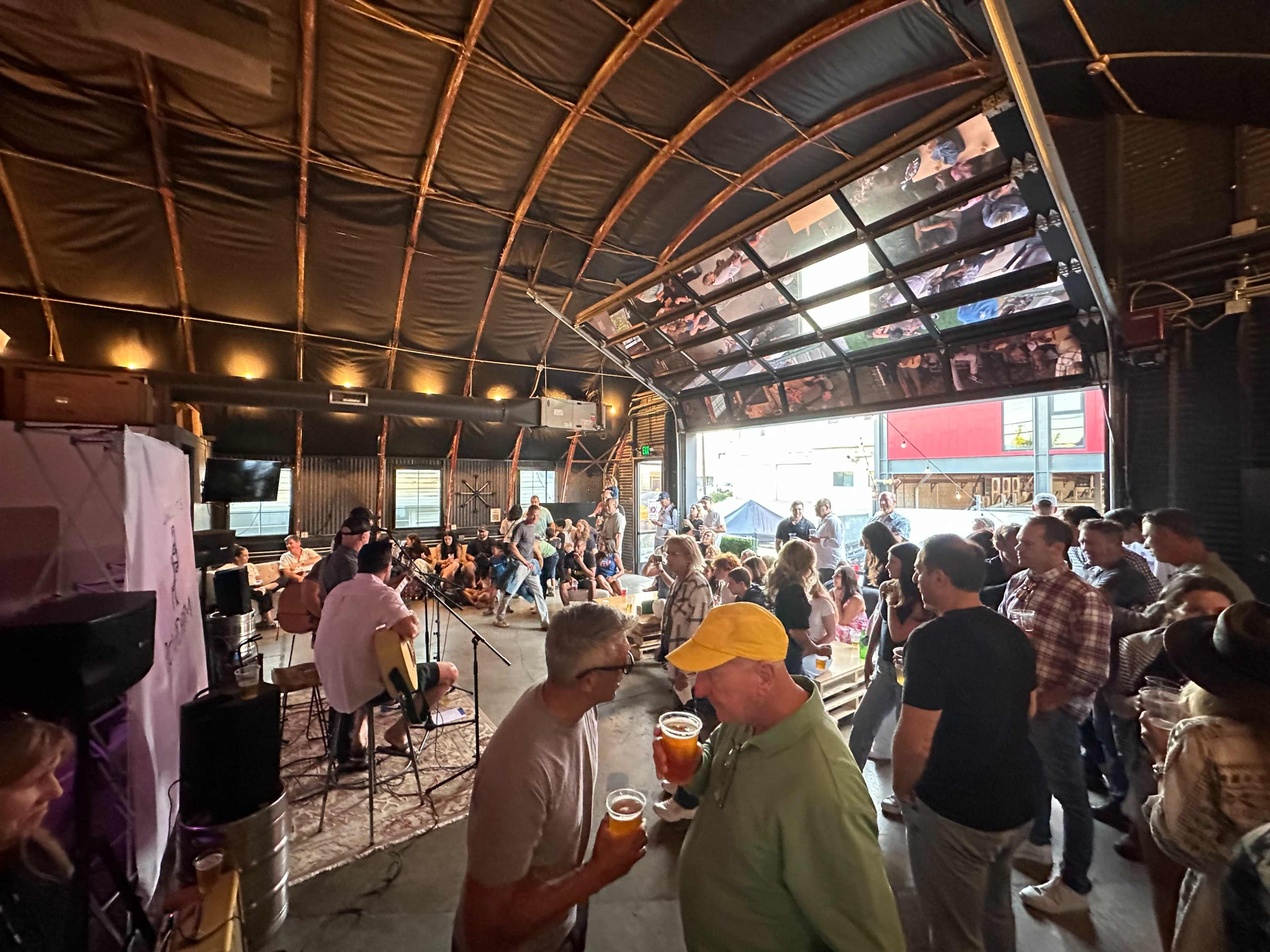 A crowd gathers in a warehouse setting, enjoying live music while holding drinks and socializing.