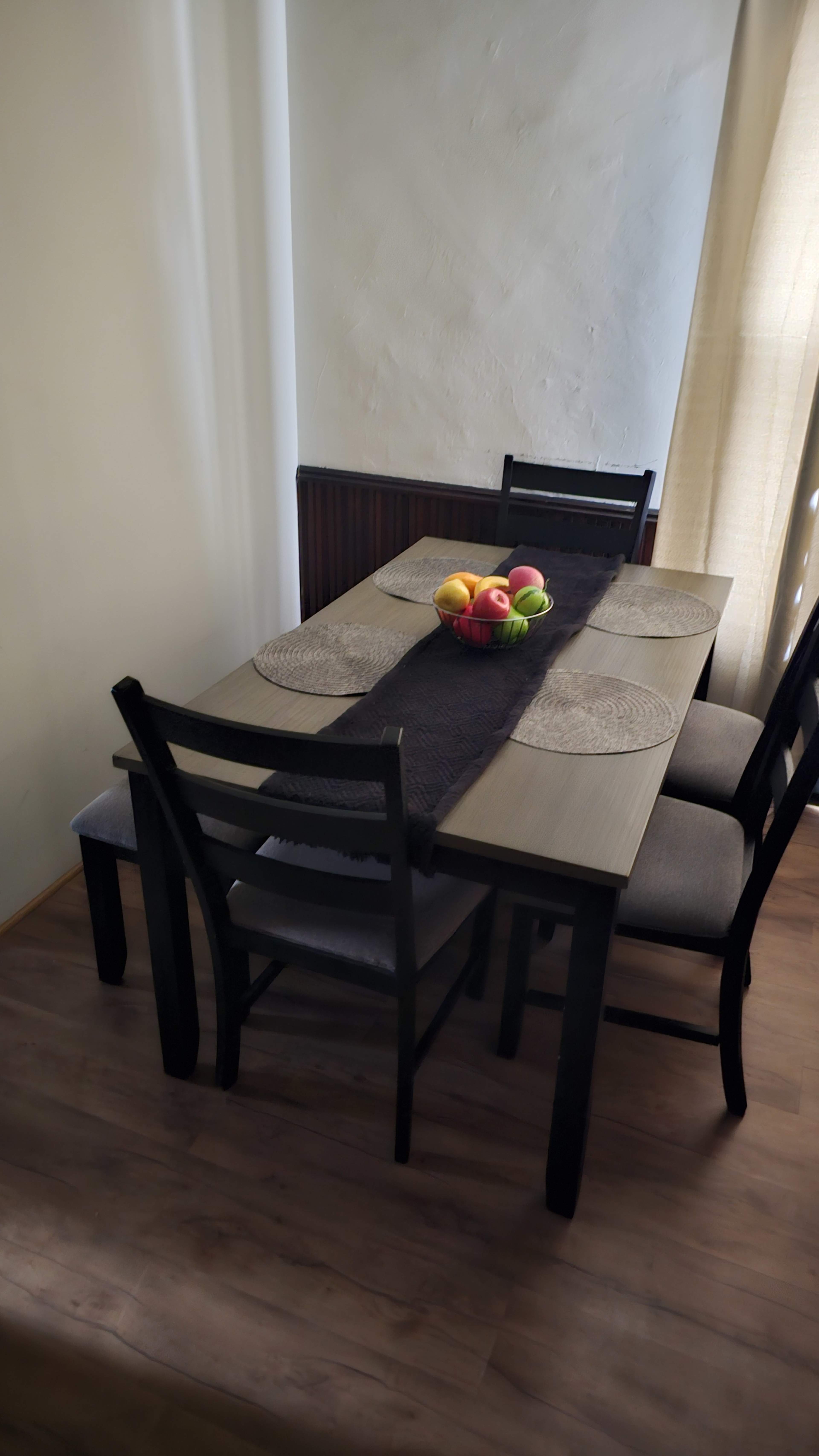 A wooden dining table with a central fruit bowl is surrounded by four chairs in a well-lit corner of a room.